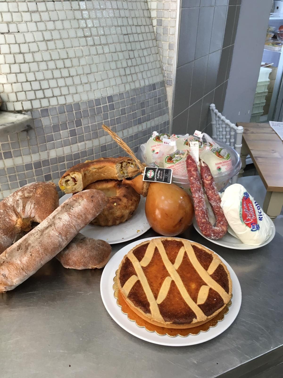 Su un bancone di metallo è disposto un assortimento di cibi, tra cui una torta salata, pane rustico, salumi e vari formaggi italiani.