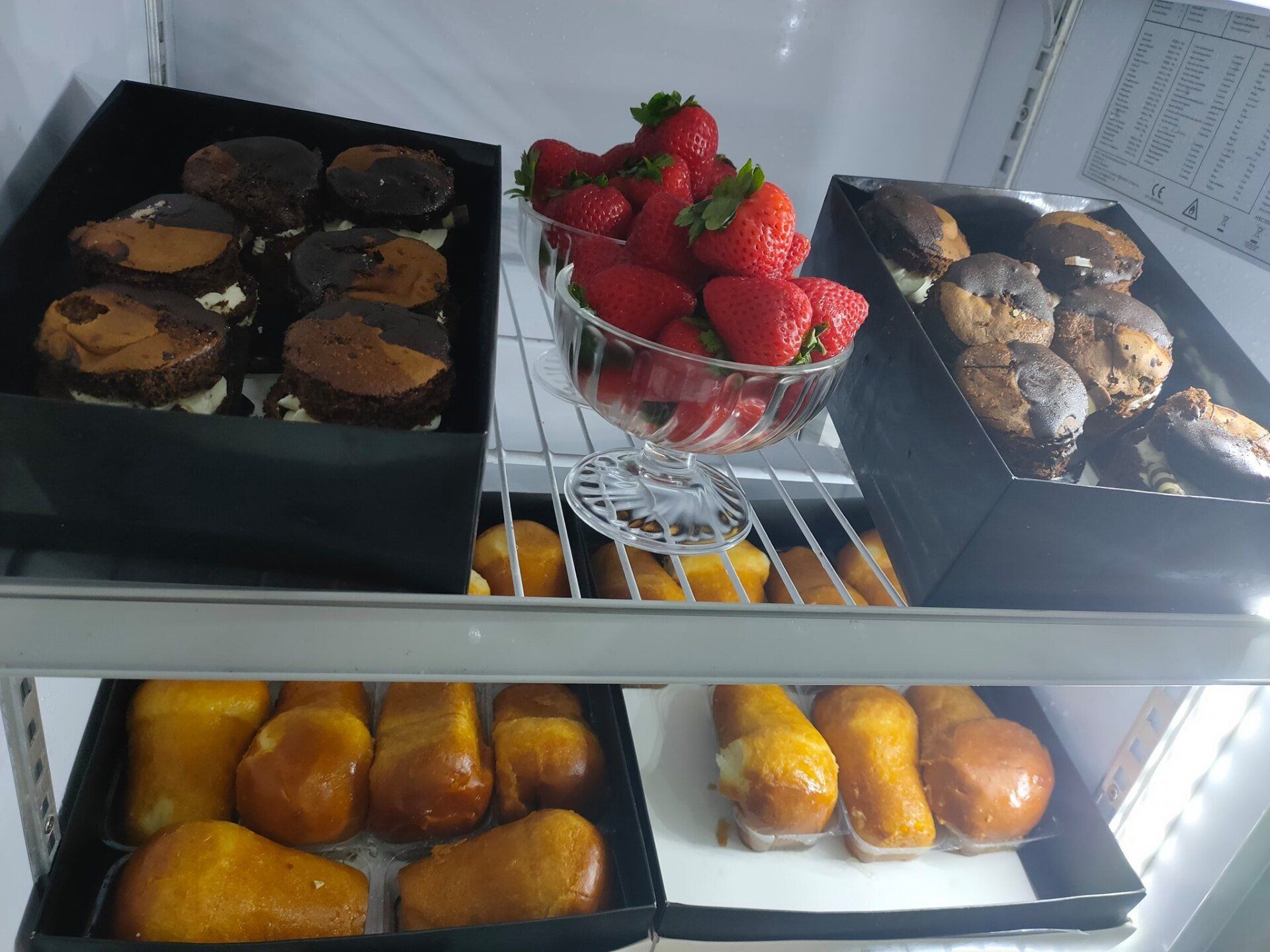 Due ripiani refrigerati contenenti scatole di pasticcini ricoperti di cioccolato, una ciotola di vetro con fragole e babà al rum dorati.