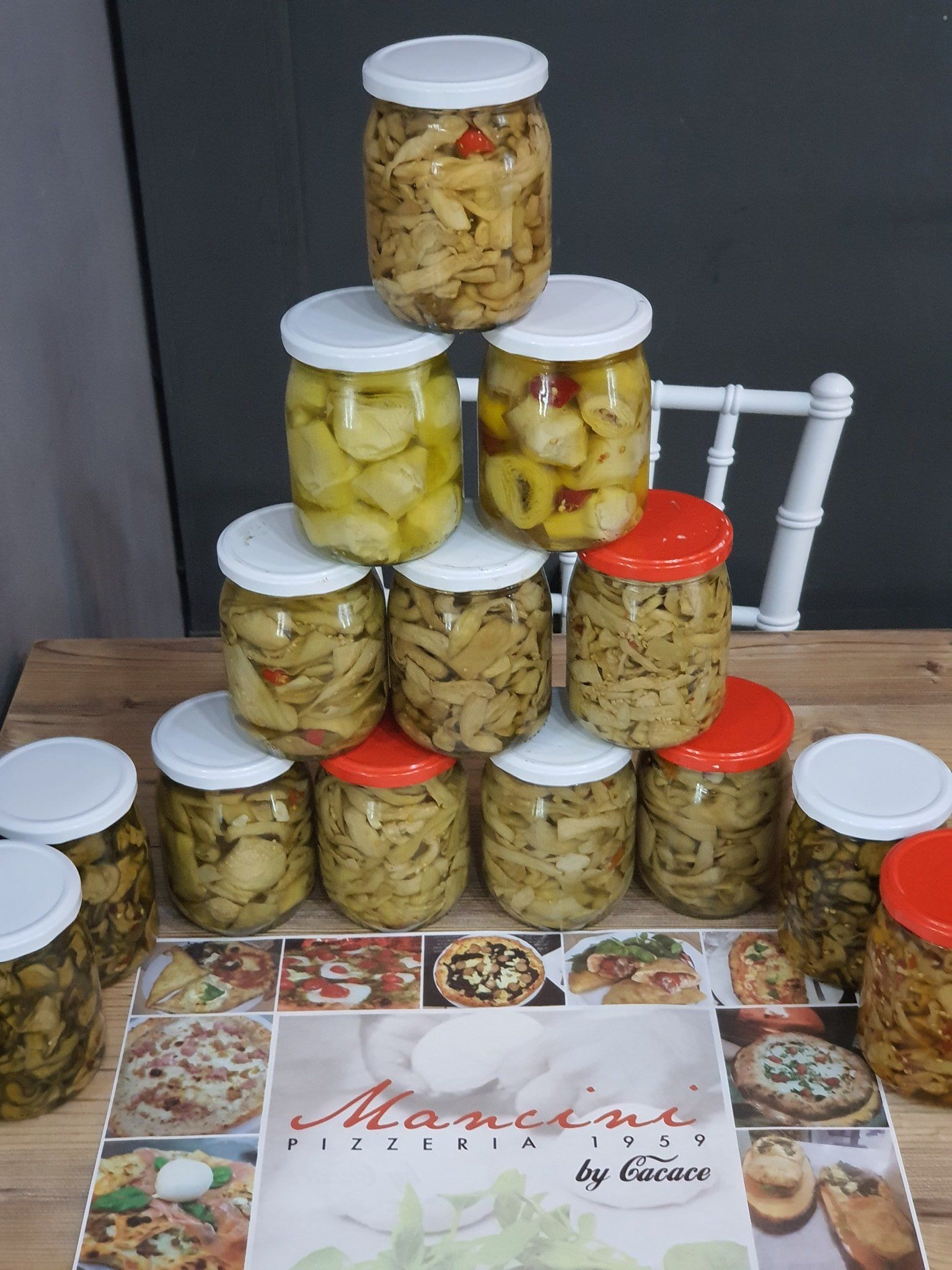 Su un tavolo è posizionata una piramide di barattoli di vetro pieni di verdure sott'aceto, accanto alla quale si trova una ricetta per le pizze Mancini.