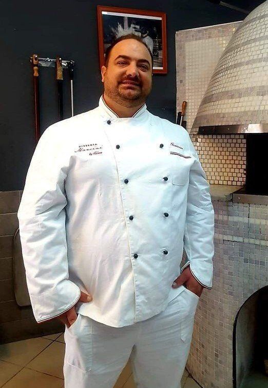 Uno chef in uniforme bianca sorride nella cucina di un ristorante, accanto a un forno in pietra rivestito di piastrelle.