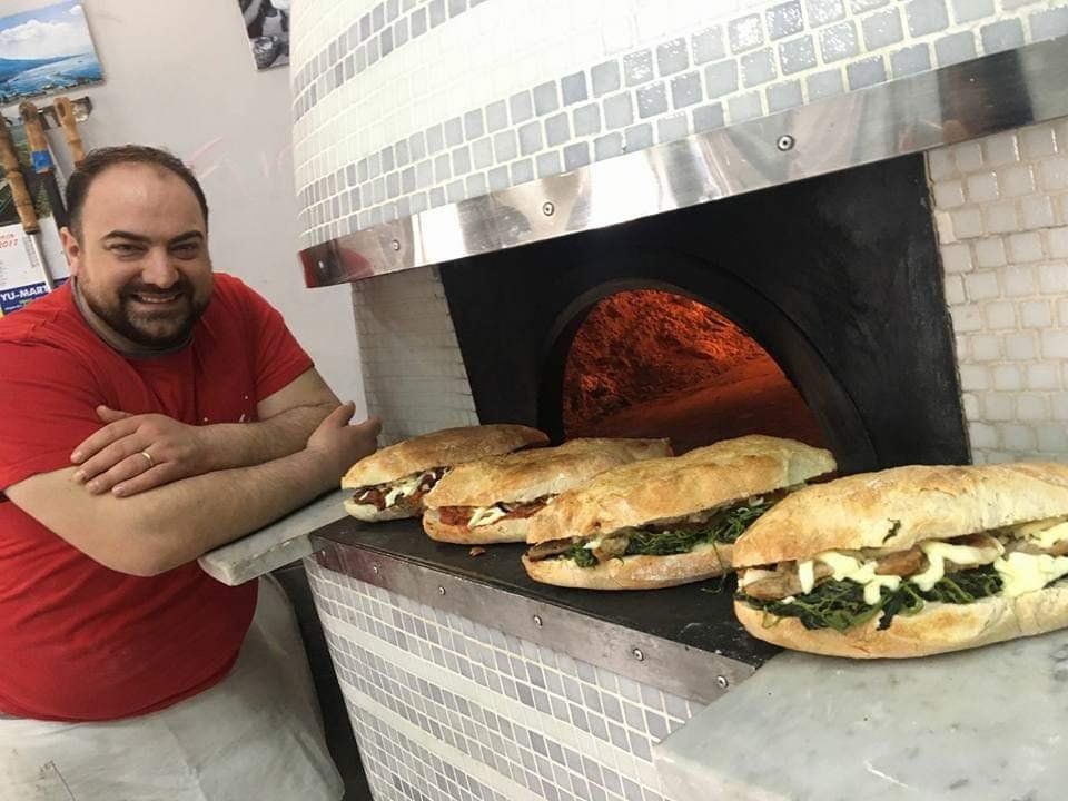Uno chef sorridente con una camicia rossa è appoggiato a un forno a legna, davanti al quale sono esposti quattro panini già pronti.