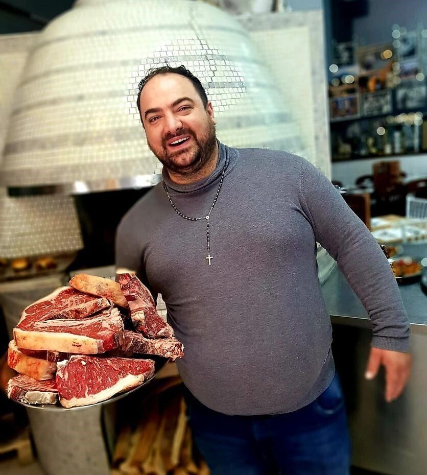 Un uomo sorridente in un ristorante, con in mano un grande vassoio di bistecche crude davanti a un tradizionale forno per pizza.