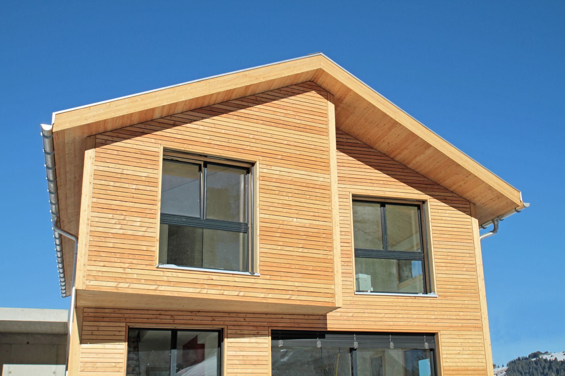A modern wooden house with large windows and a sloped roof, set against a clear blue sky.