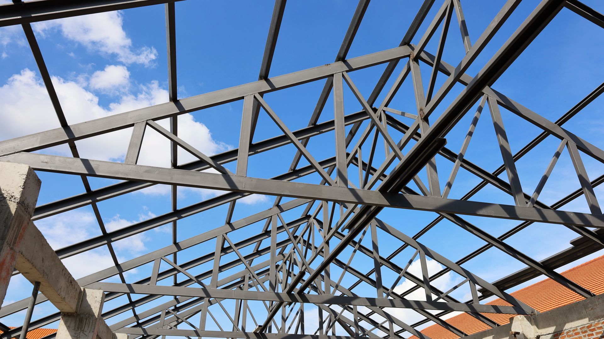 Steel roof truss framework under construction with blue sky and clouds overhead. Steel roof truss framework under construction with blue sky and clouds overhead.