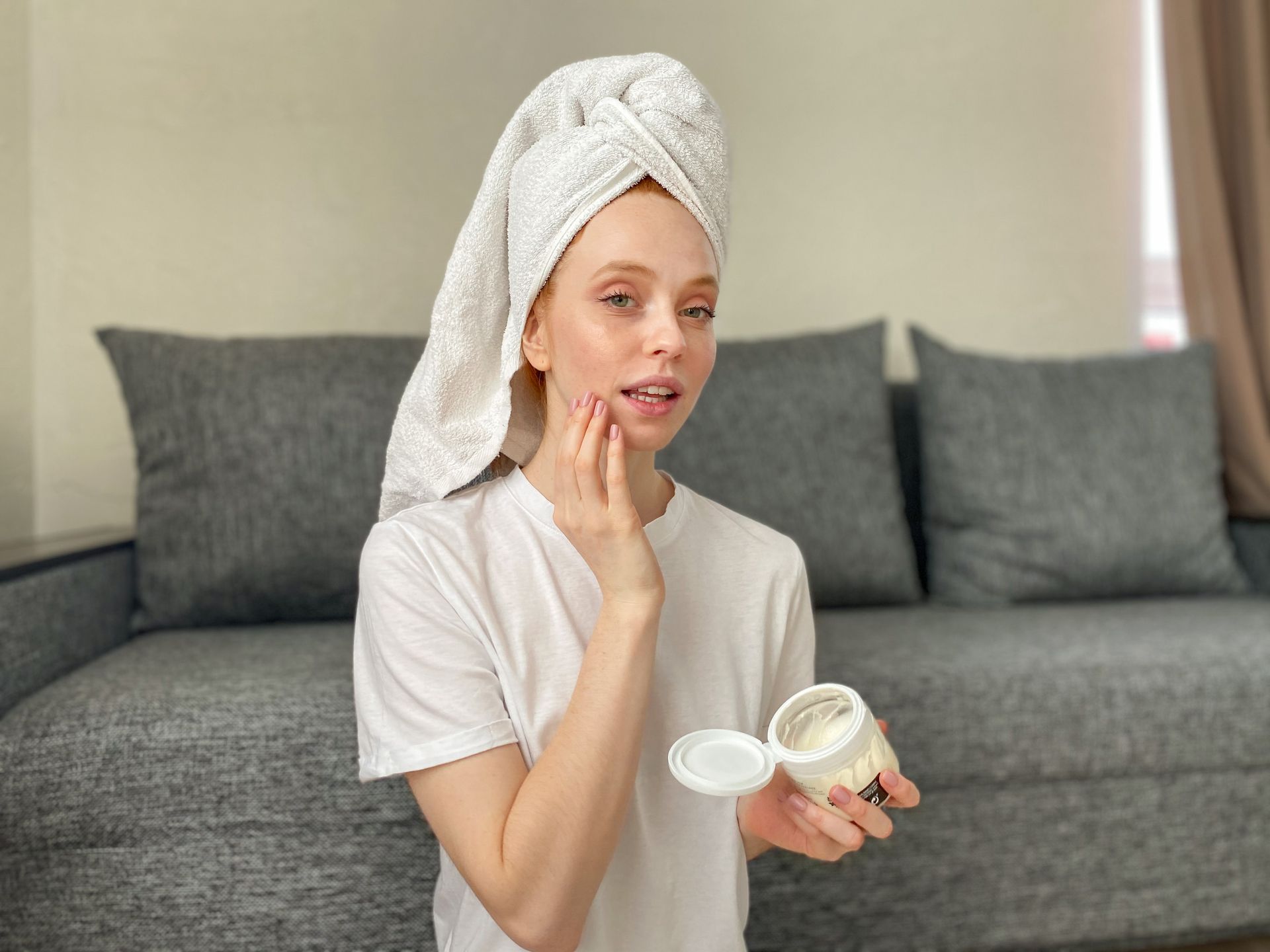 A woman with a towel wrapped around her head is applying a cream to her face.