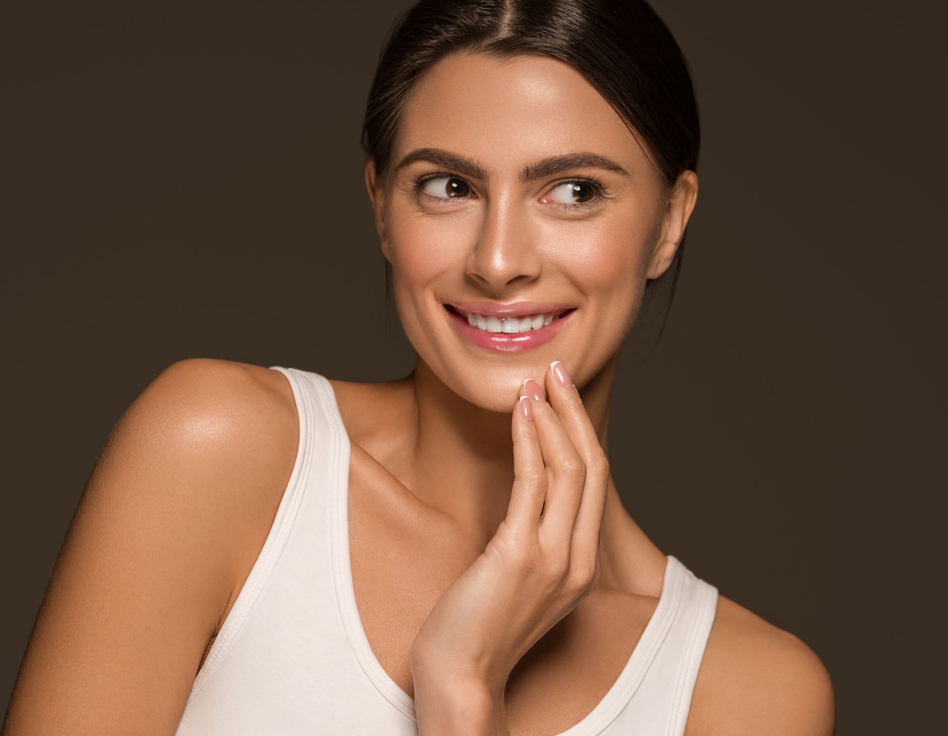 Woman with tan skin and dark hair in a white tank top smiles, touching her face, set against a brown background.