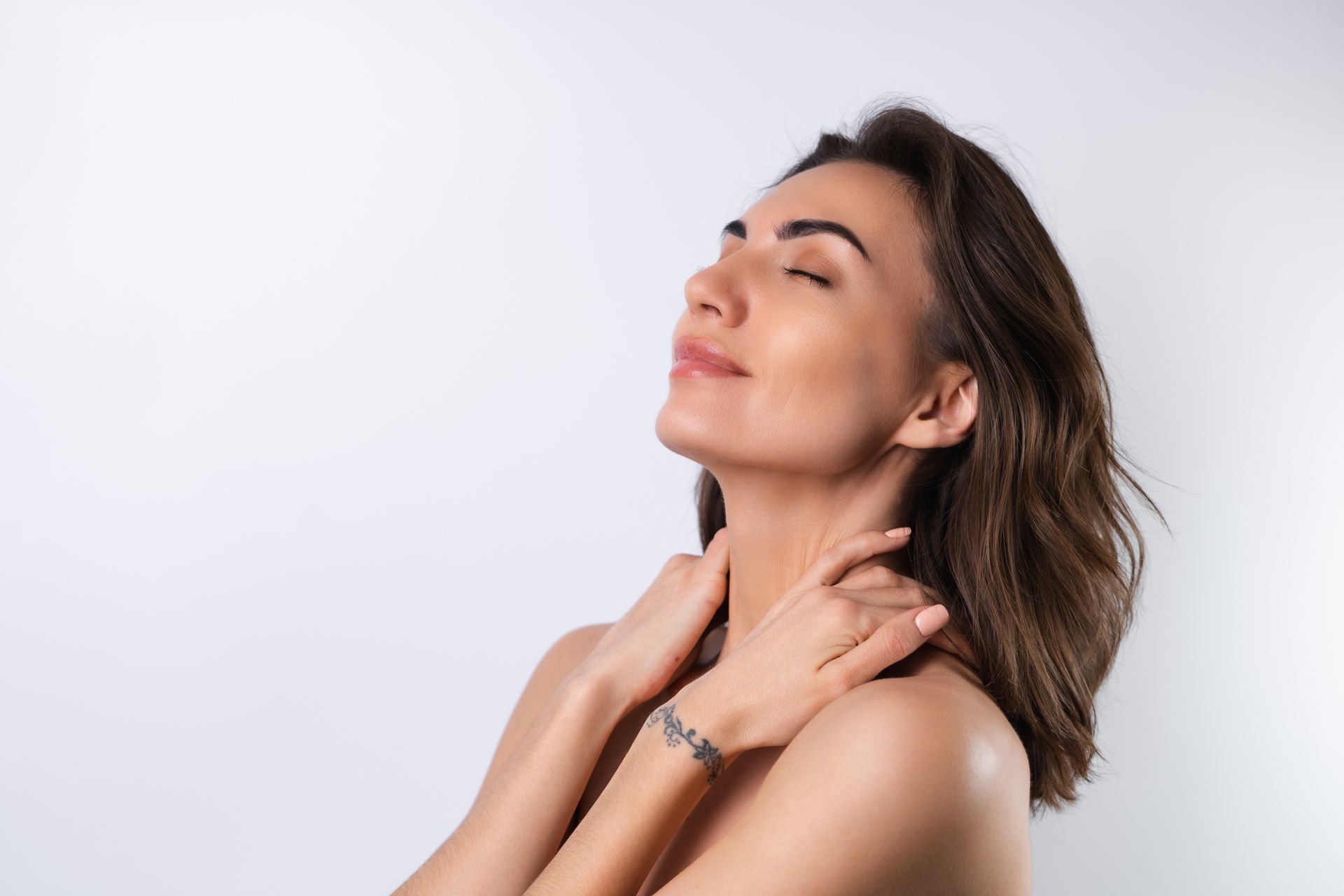 A person with shoulder-length brown hair, eyes closed, gently touching their neck against a light gray background.