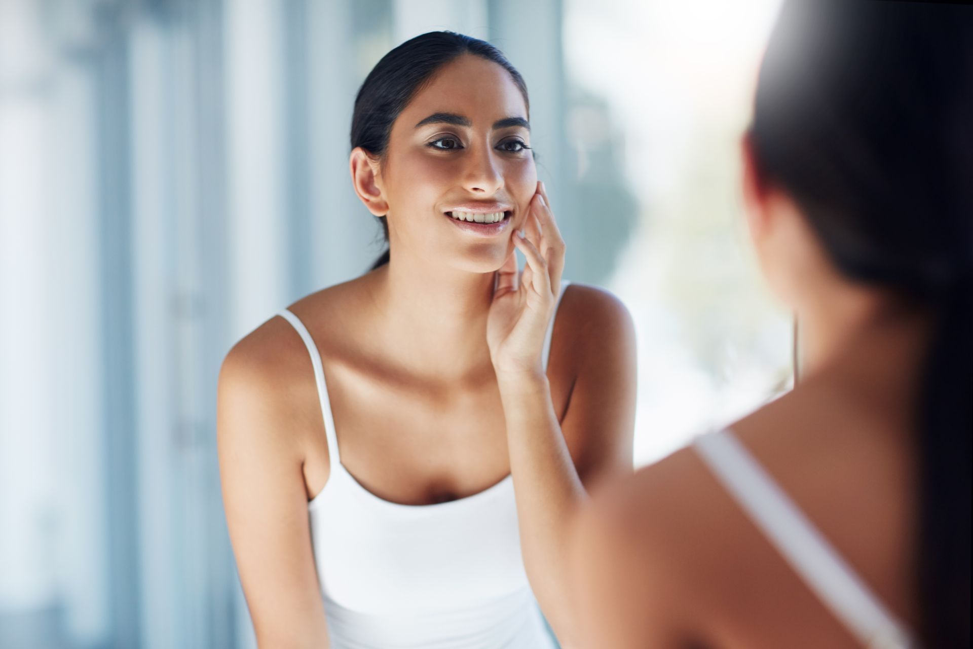 A person in a white camisole examines their skin in a mirror with a light-filled background.
