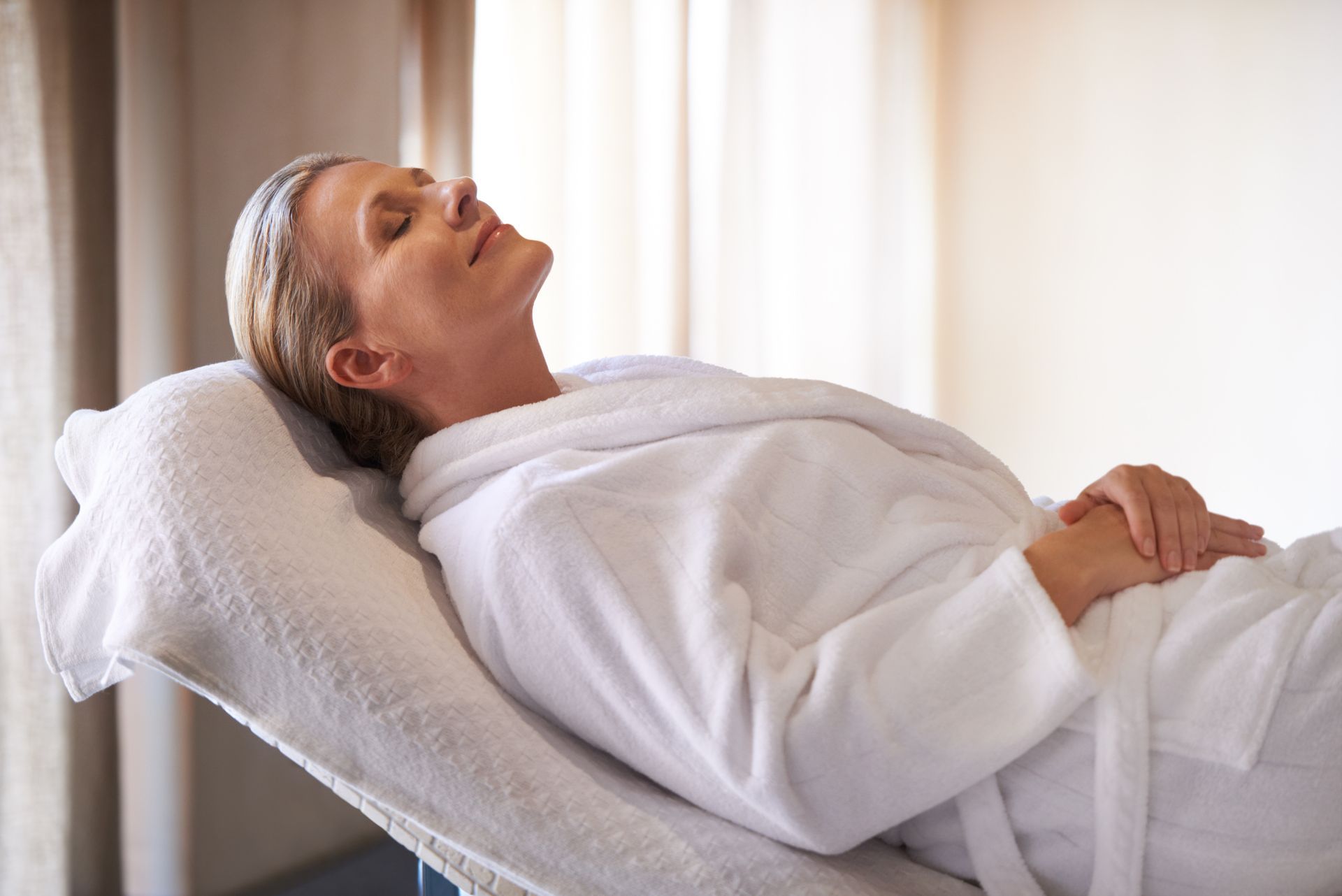 A person wearing a white bathrobe rests with their eyes closed on a reclined spa chair.