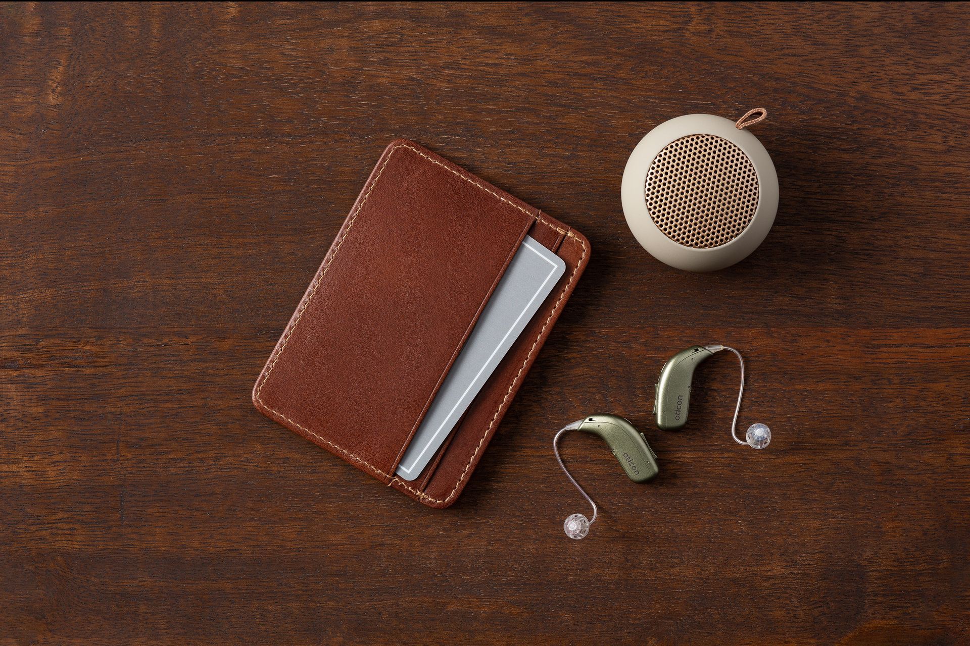 Brown wallet with card, hearing aids, and a small round case on a wooden surface.