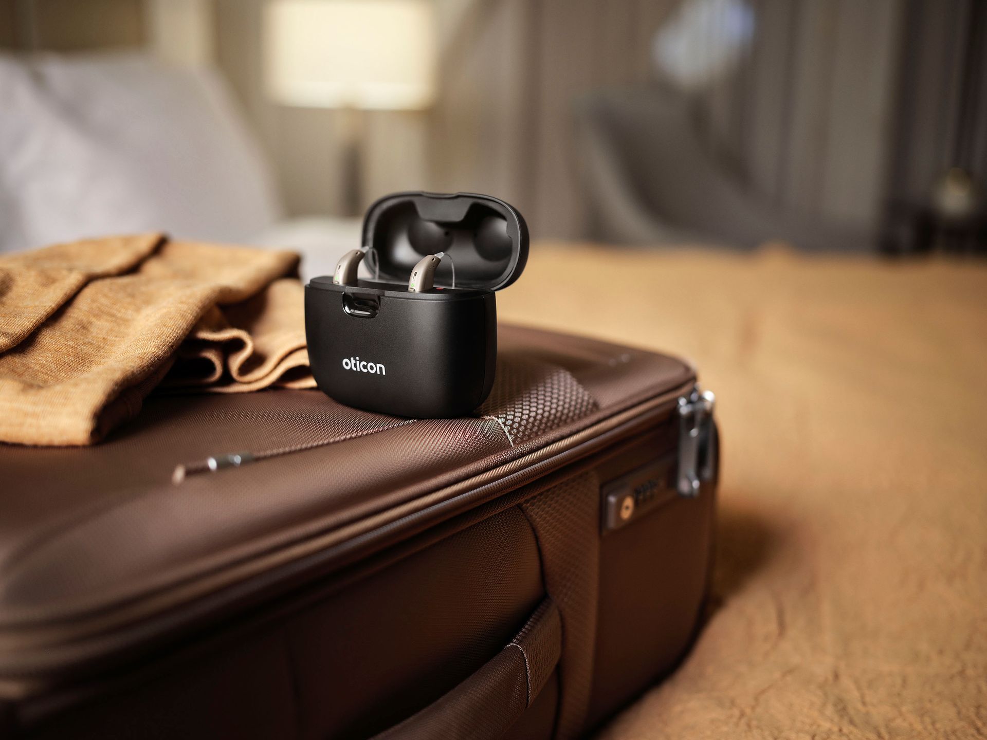 Black hearing aid charging case on a brown suitcase with a beige cloth, indoors.