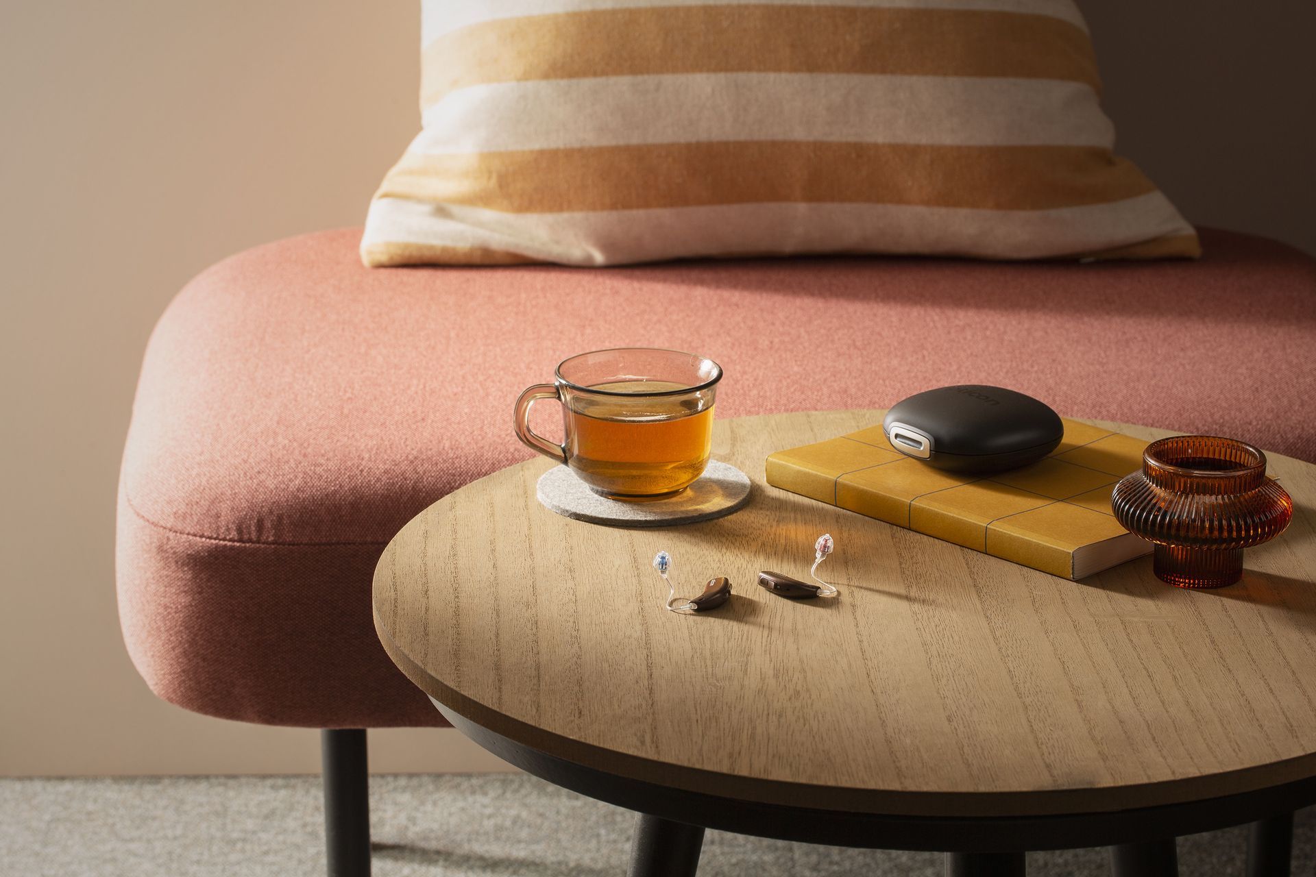 A cup of tea and a notebook on a wooden table, next to a small upholstered pink bench and striped pillow.