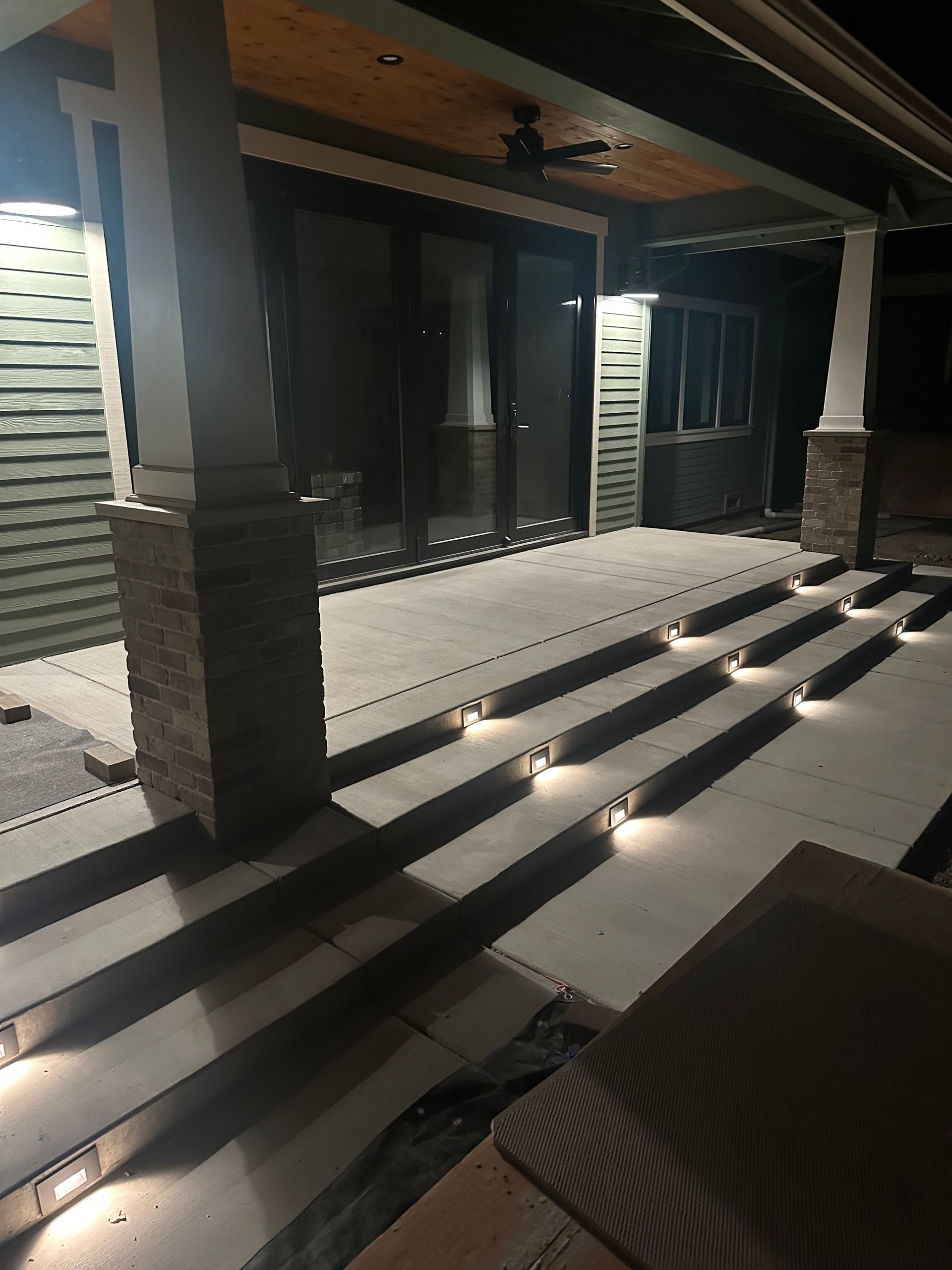 Nighttime covered porch with concrete steps, dark columns, and lit ceiling lights.