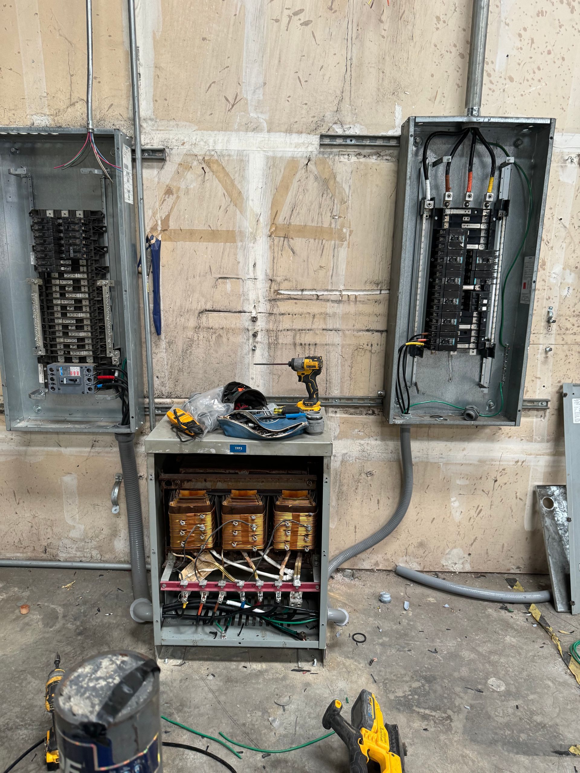Open electrical panels and wiring in a concrete utility room, with tools and parts on the floor.