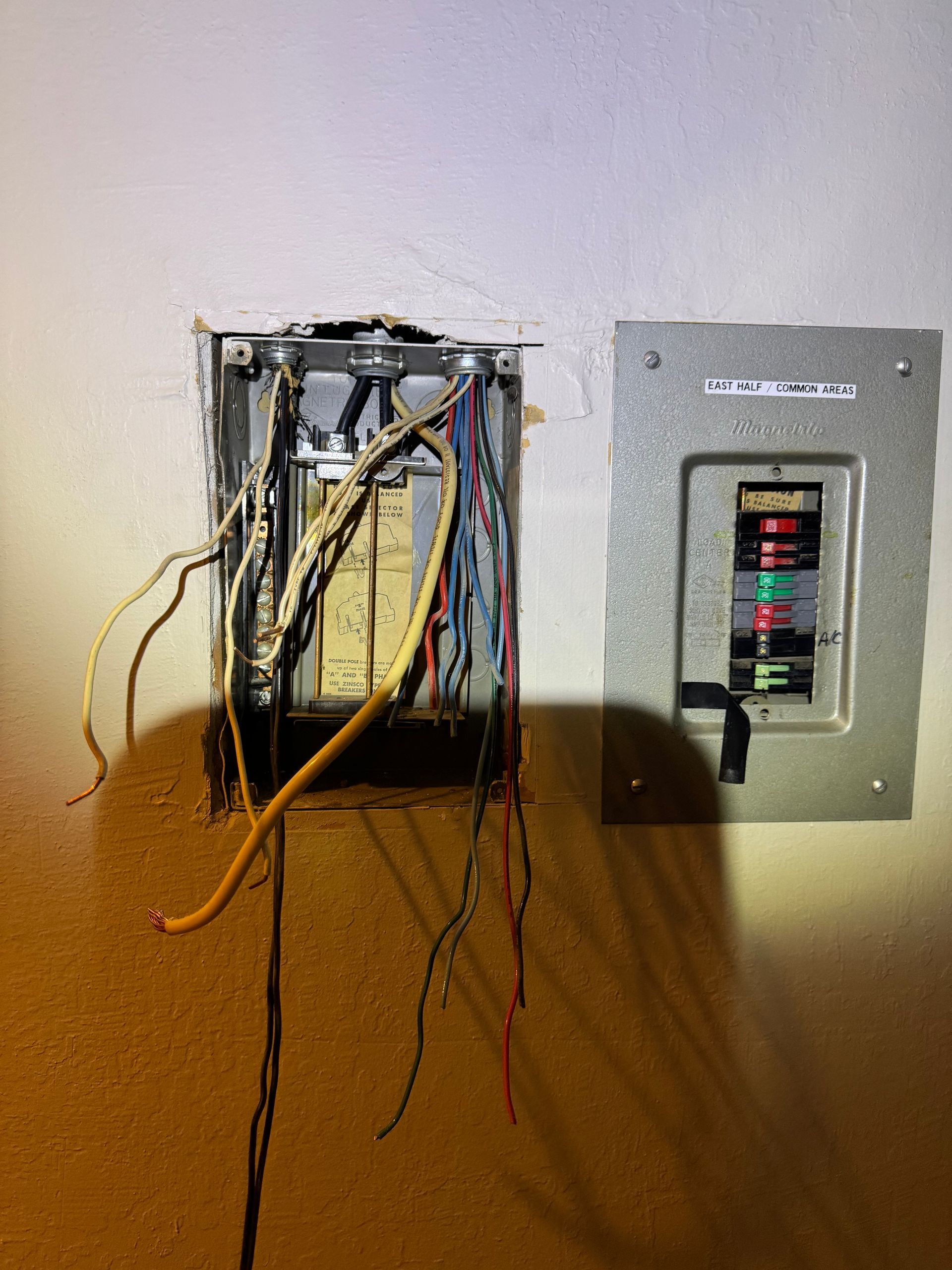Exposed electrical wiring beside a breaker panel in a wall opening, with loose cables hanging out.