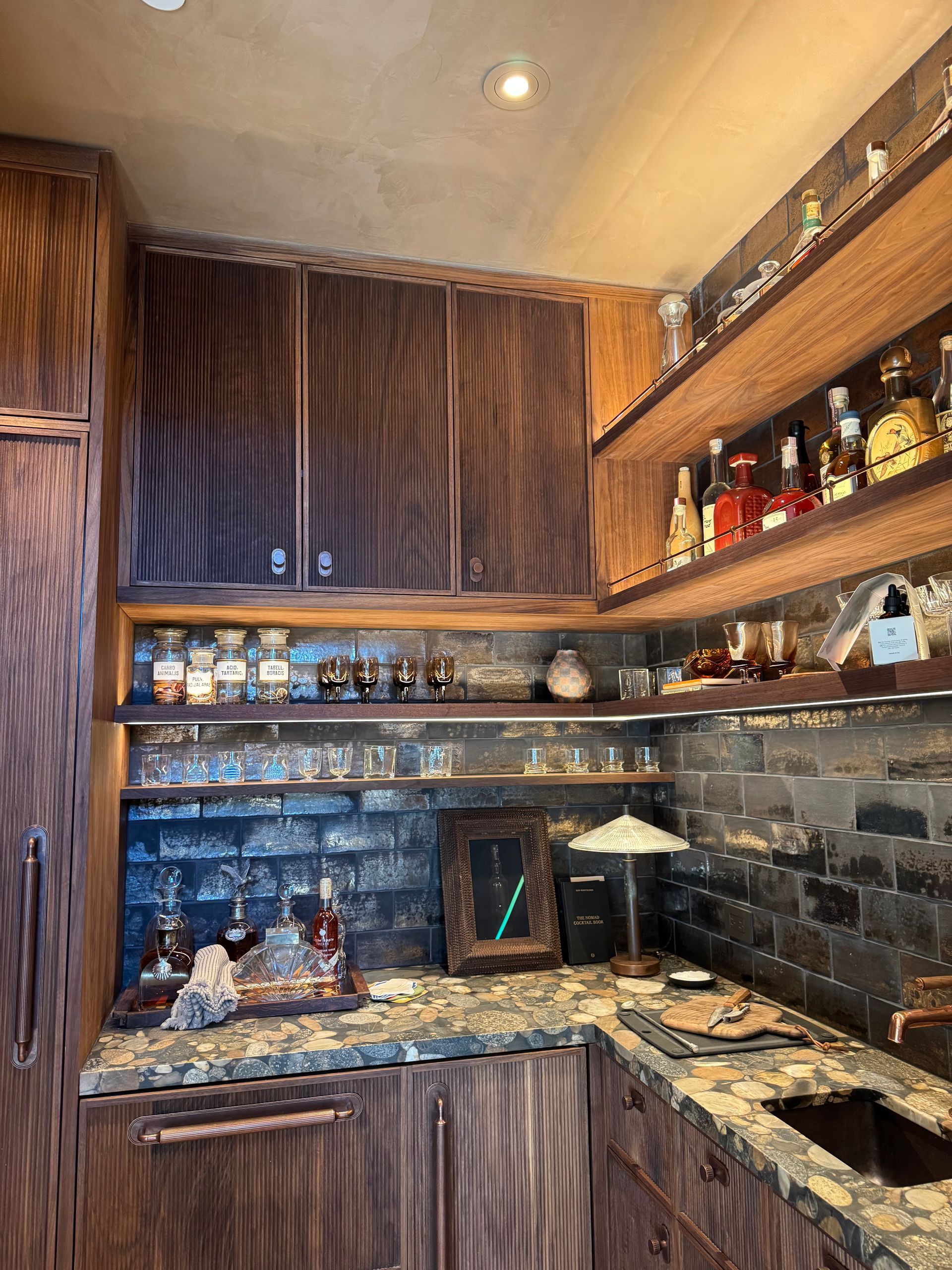 Compact kitchen bar with wood cabinets, stone countertops, and shelves of bottles and glassware