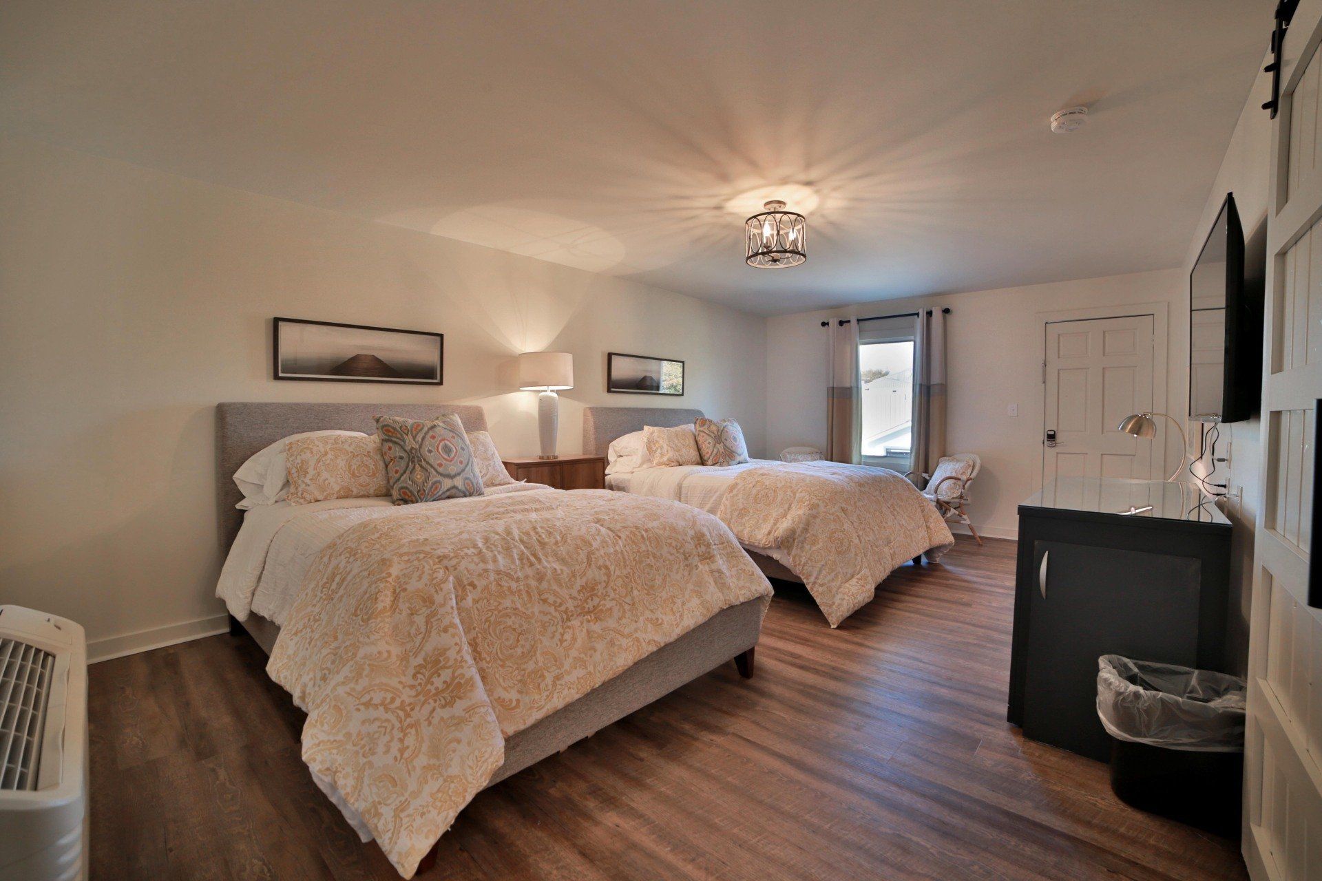 A bright, clean hotel room with two beds covered in fluffy white blankets, a small TV, and a wood floor.