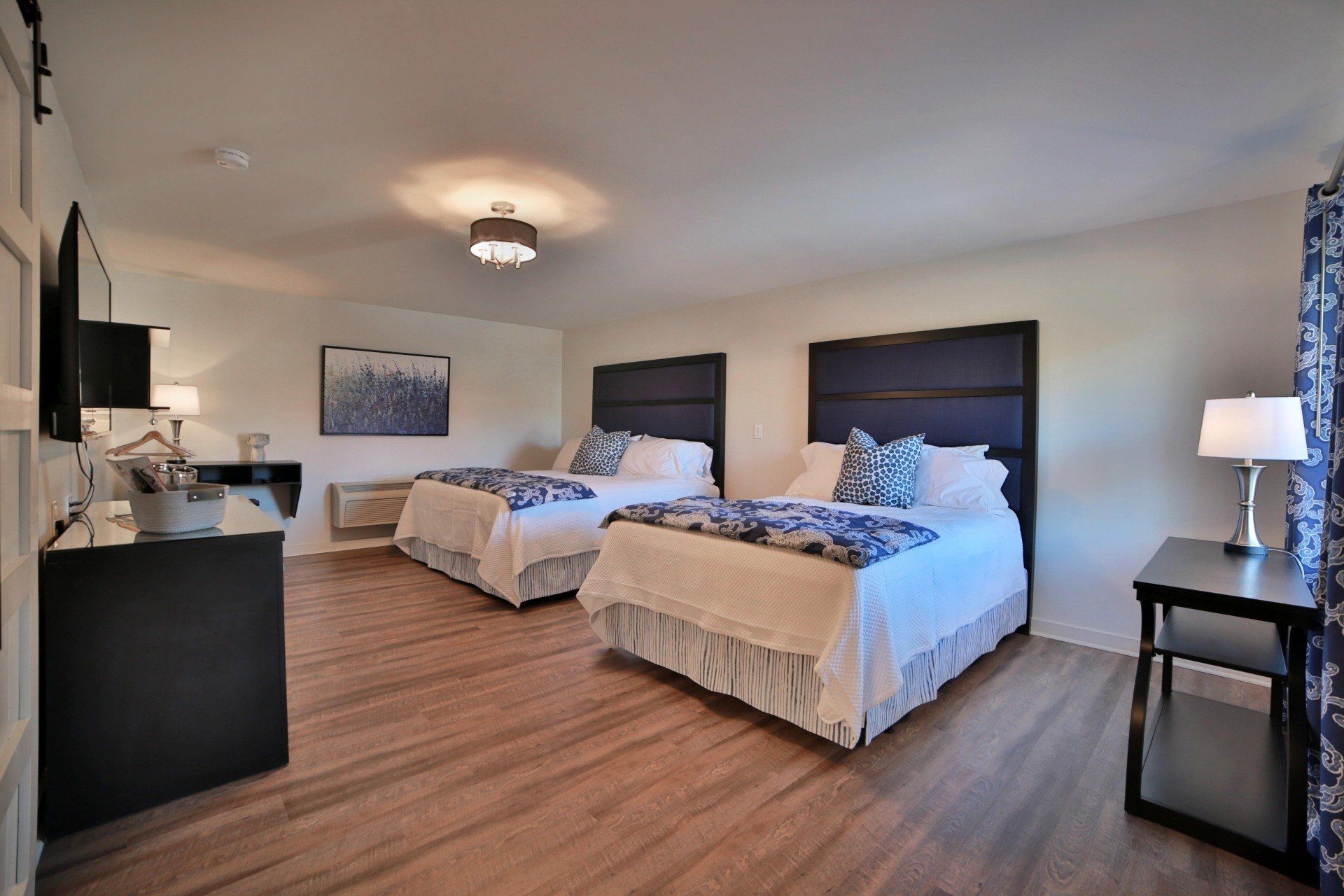 A hotel room with two double beds, each with blue accents, set against a white wall. A TV, desk, and lamp are also present.
