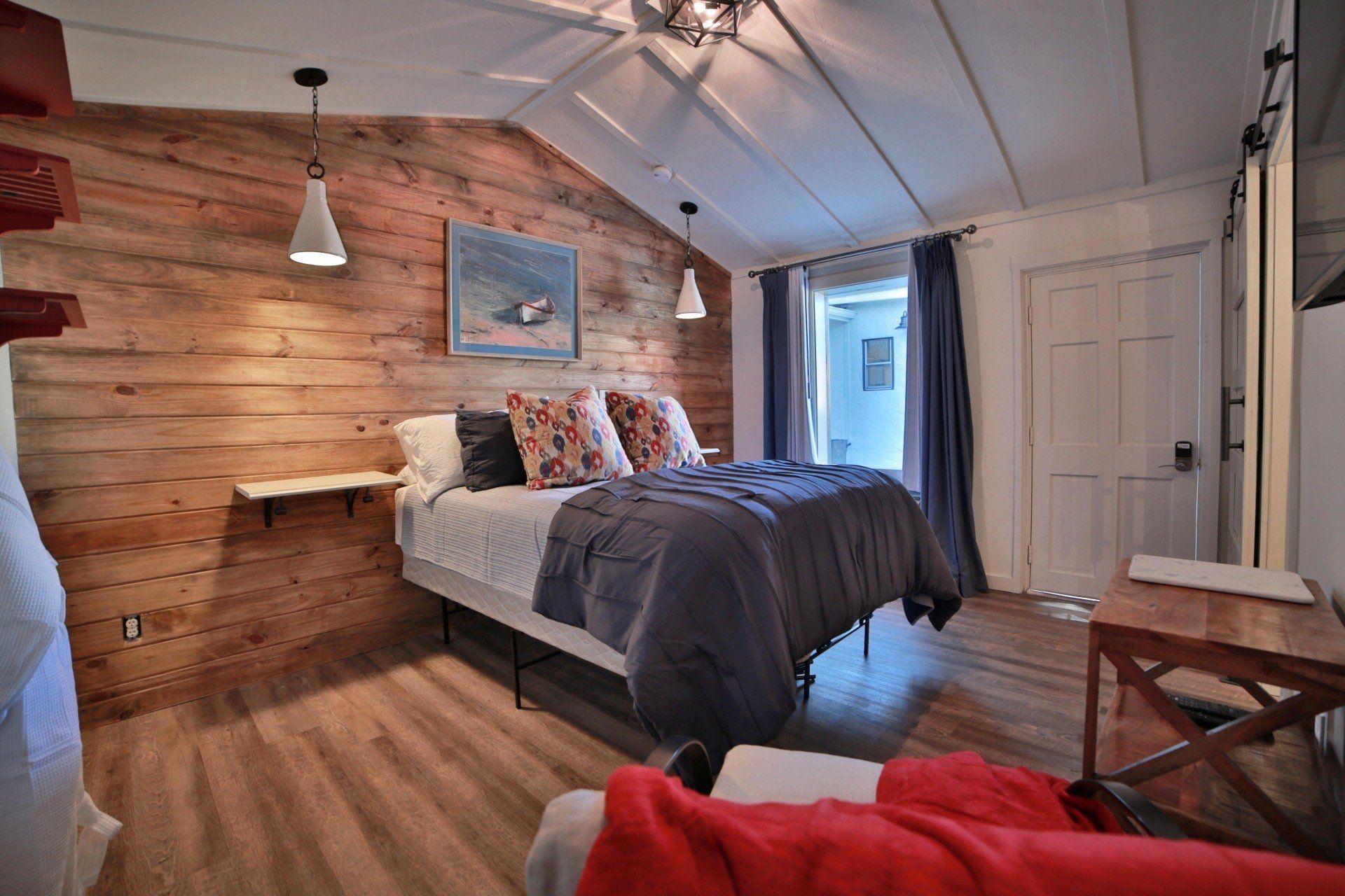 A bedroom with a rustic wood wall, a bed with pillows and a blue comforter, and a door to the outside.