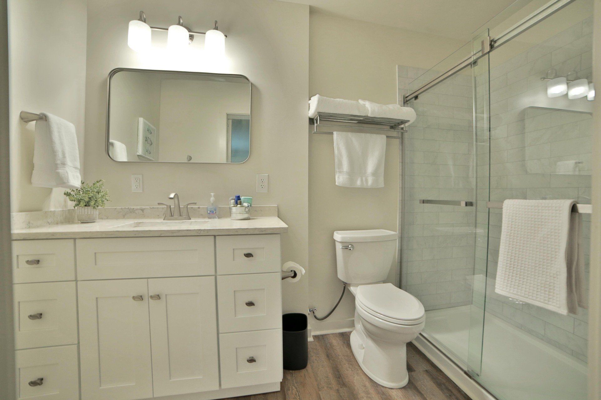 White bathroom with a vanity, toilet, and shower.  A mirror hangs above the vanity.  The floor has wood-like tiles.
