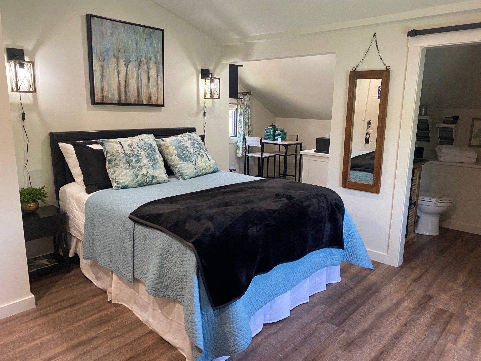 Bedroom with a bed, art, mirror, and an adjacent room with a table and a bathroom visible.