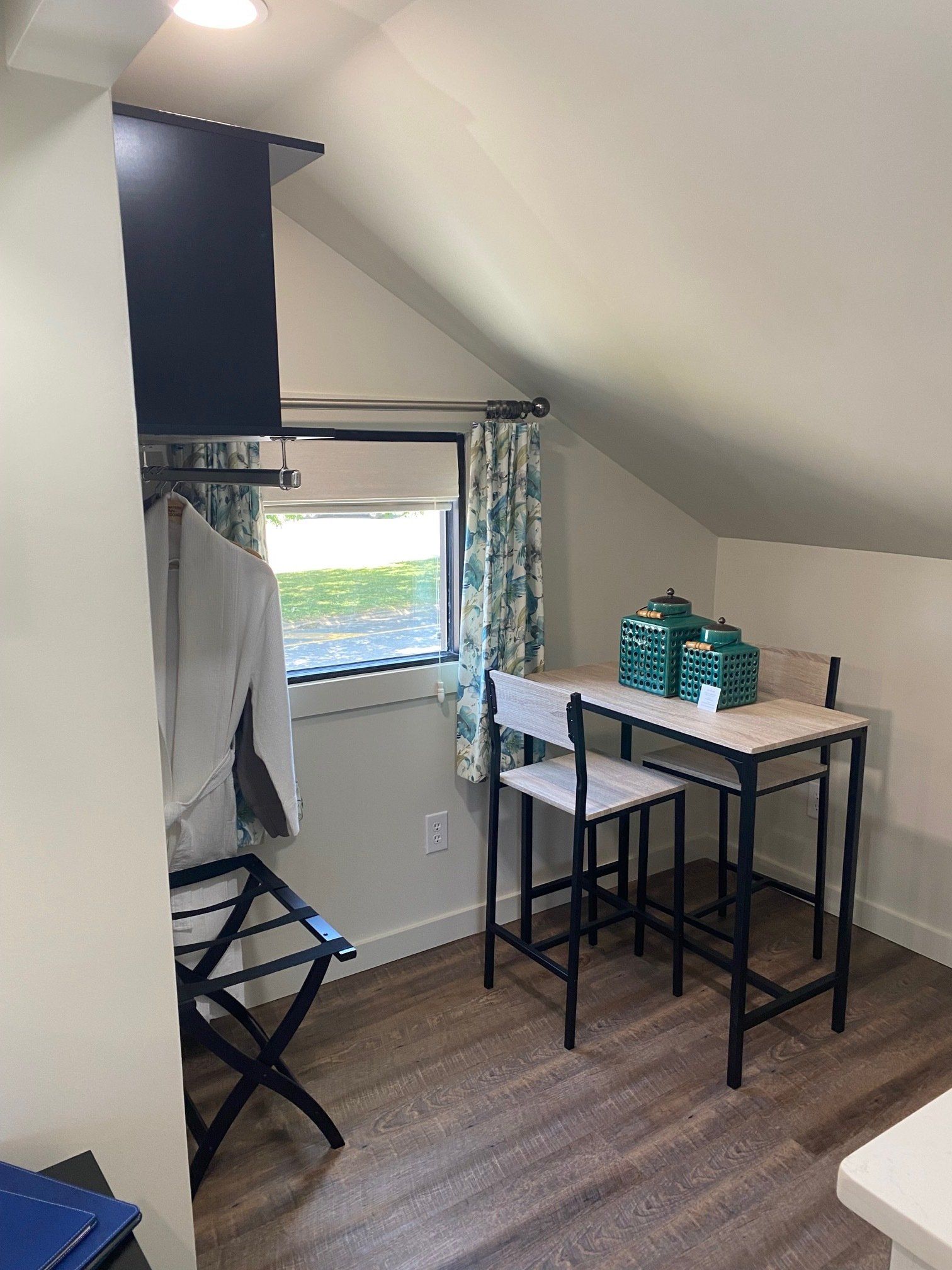 Small room with sloped ceiling. A dining set, window with curtains, clothing rack, and suitcase stand are visible.