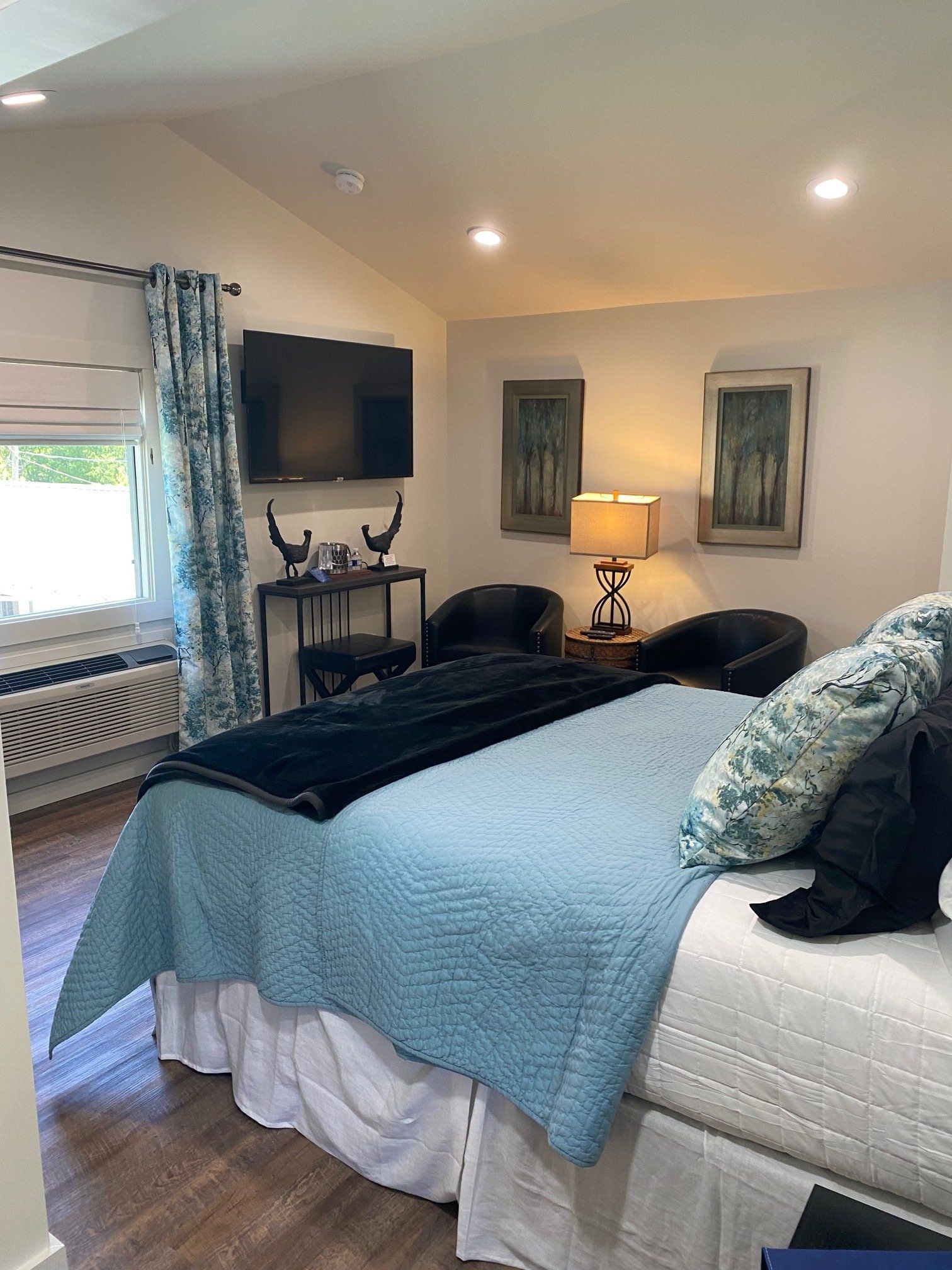 A bedroom with a bed, a TV, and seating. The bed has blue bedding, and the room has natural light.