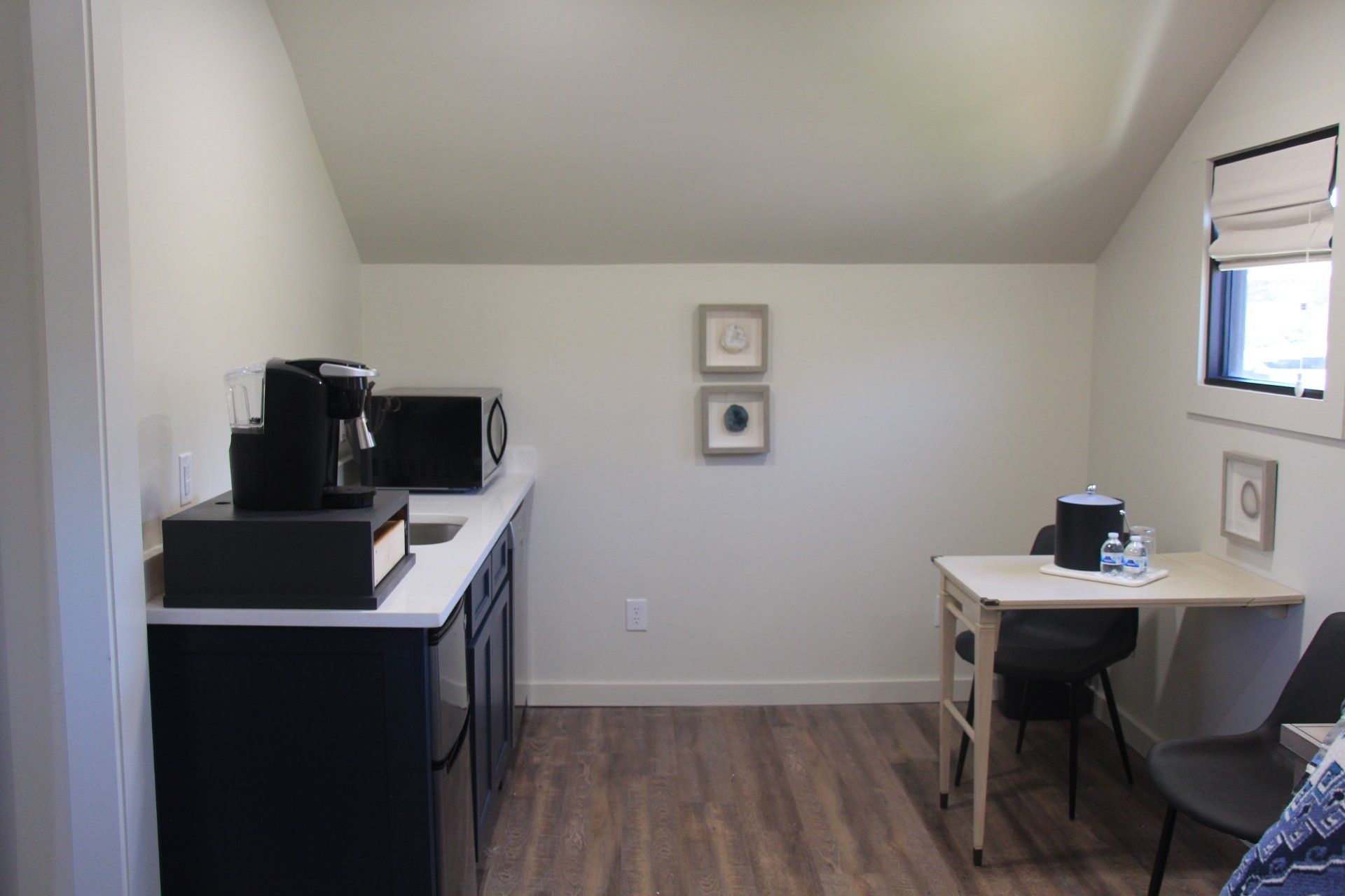Small kitchenette with a microwave, coffee maker, and table with chairs. Light-colored walls and wooden floor.