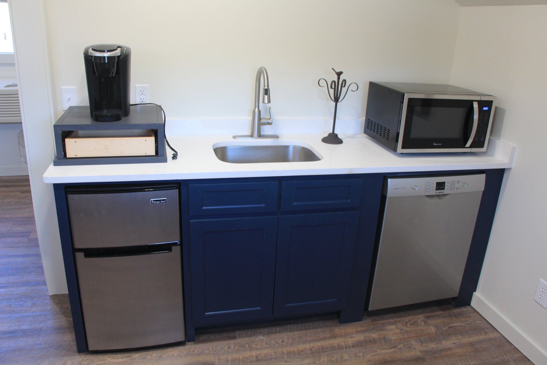 A compact kitchen with a stainless steel mini-fridge, sink, dishwasher, microwave, and coffee maker on a blue cabinet.