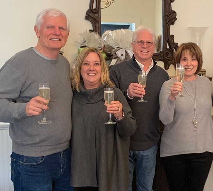 Four people in gray sweaters and jeans clink champagne flutes, smiling. Standing in front of a dark antique mirror.