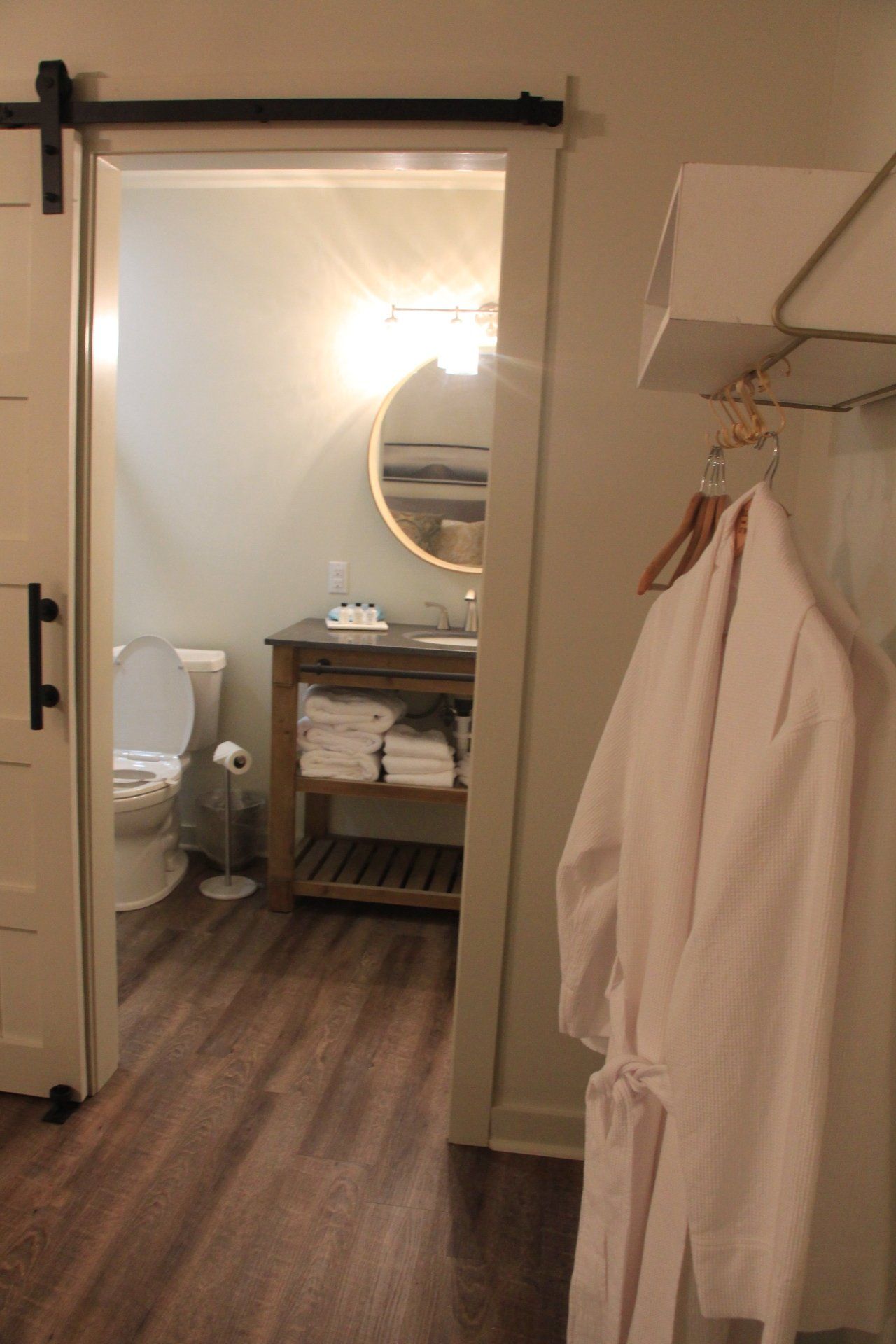 A white bathrobe hanging in a doorway leading to a bathroom with a toilet, sink, and round mirror.