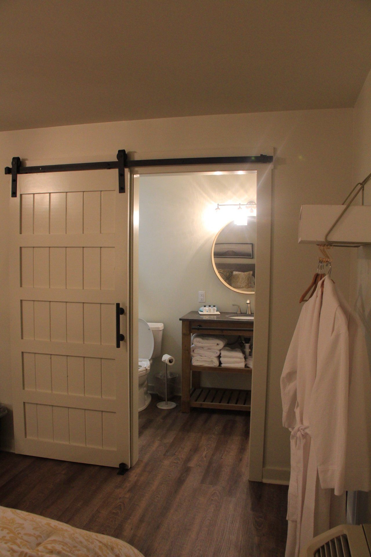 A barn door slides open to reveal a bathroom with a round mirror, vanity, and toilet.