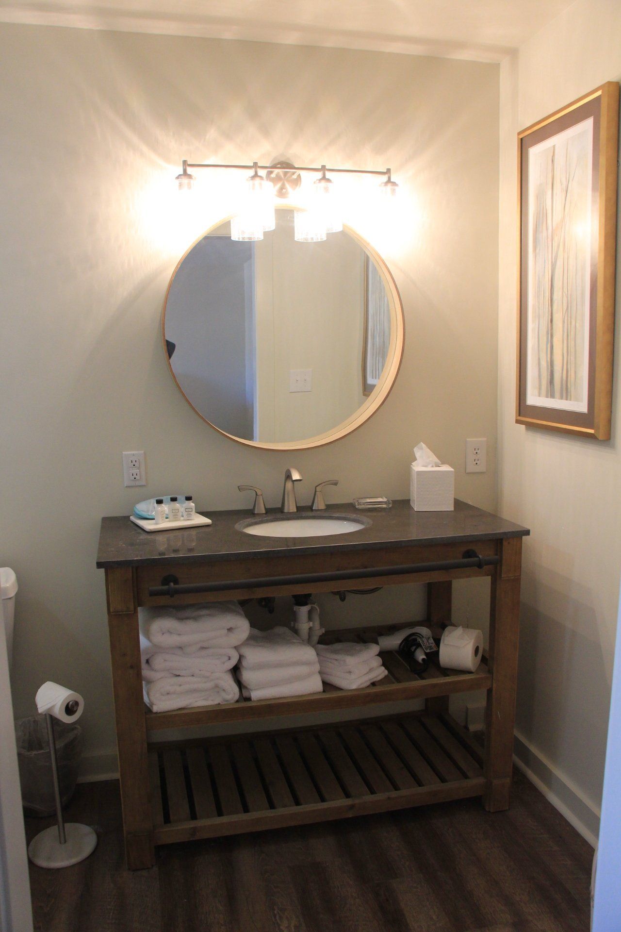 Bathroom with a round mirror, vanity with towels, and a framed art piece on a neutral wall.