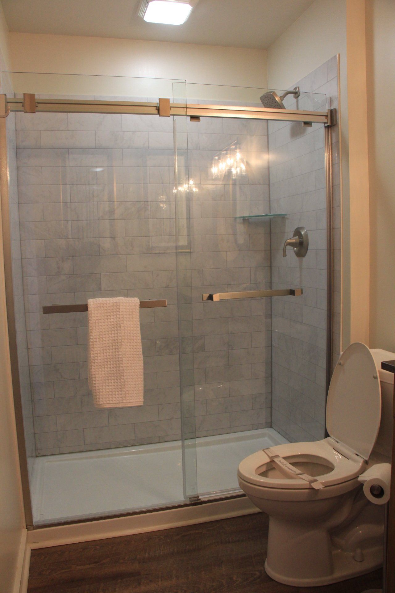 A bathroom with a glass shower, toilet, and a towel. The floor is dark wood, and the walls are light tan.