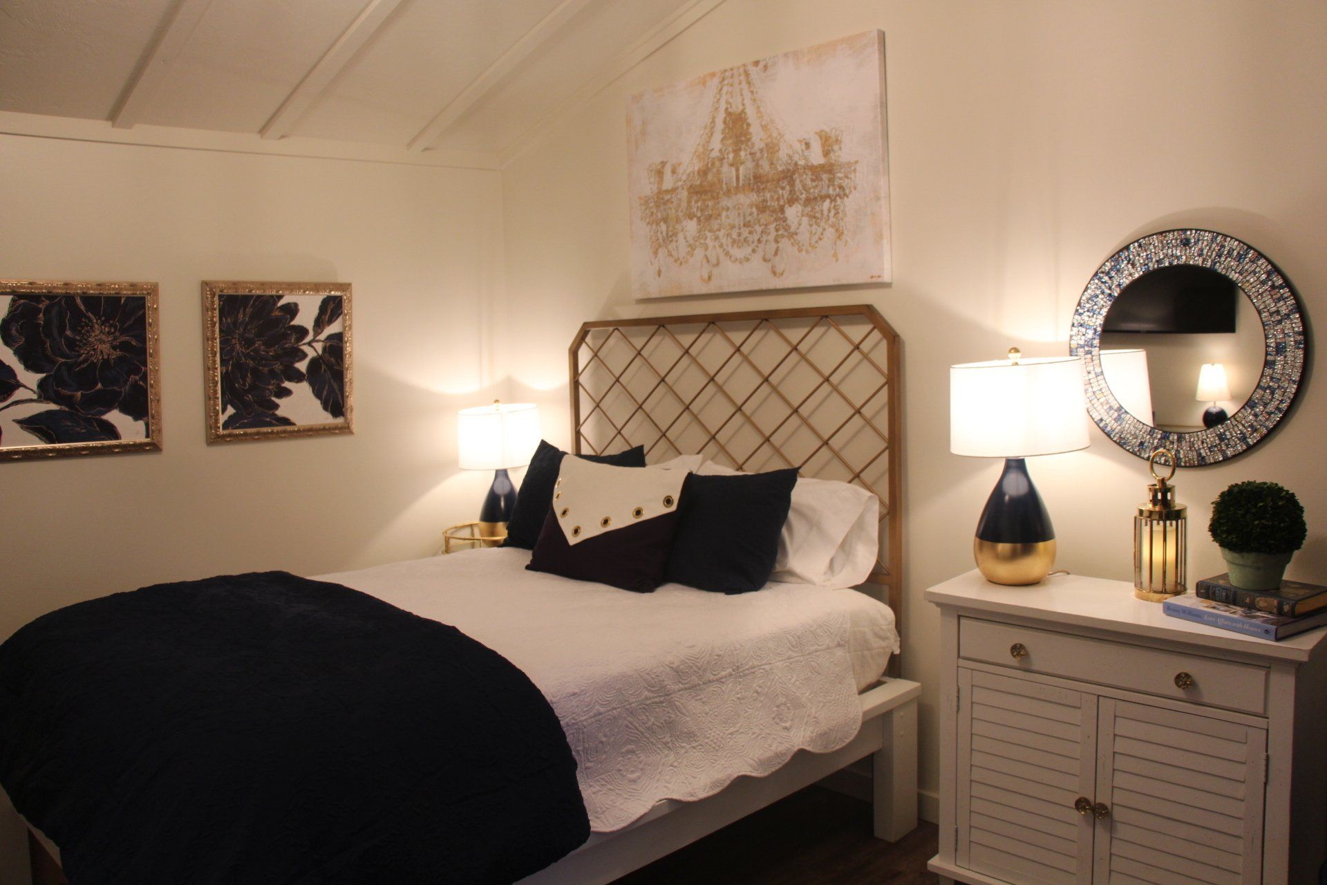 Cozy bedroom with a white bed, navy blue accents, and gold-framed art, including a chandelier-style painting.