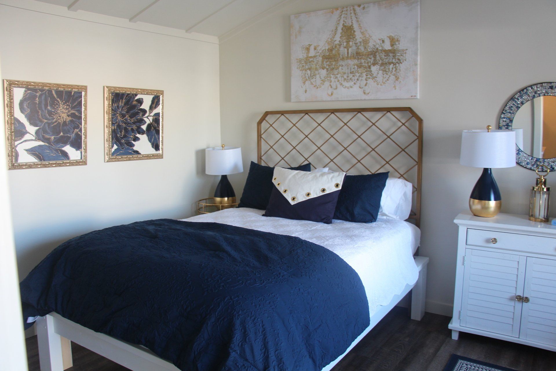 Cozy bedroom with navy and gold decor. Includes a bed, nightstand, artwork, and round mirror.