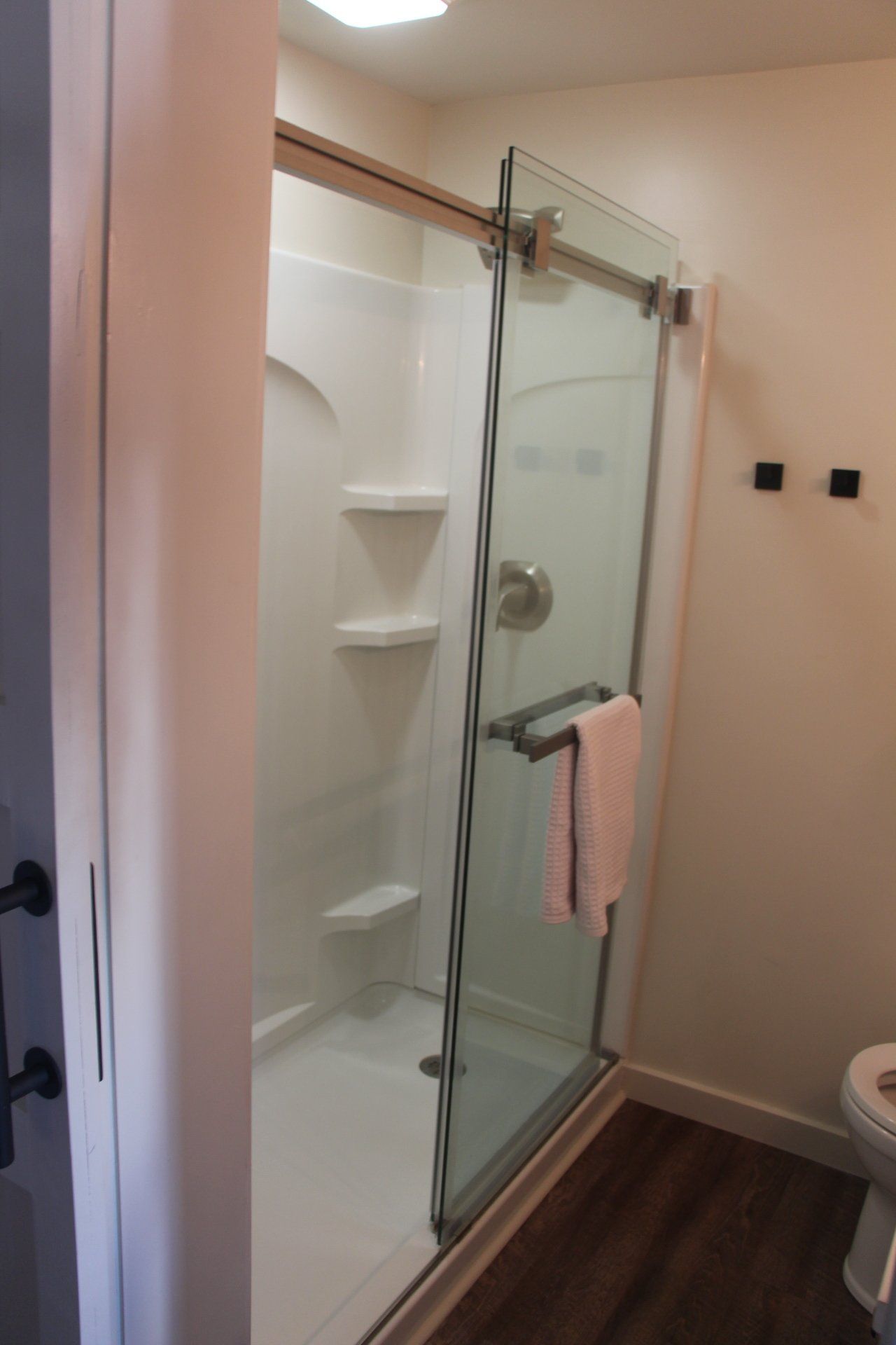A white shower with glass door and towel rack, beside a door and toilet. Brown flooring.