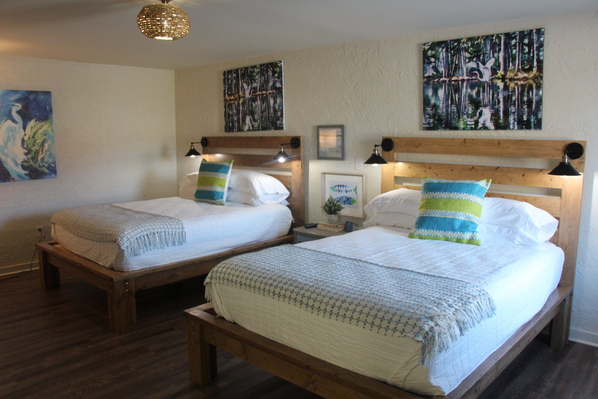 Two beds with wooden frames in a bedroom, each with a light blue, lime green, and white pillow. Artwork hangs above the beds.