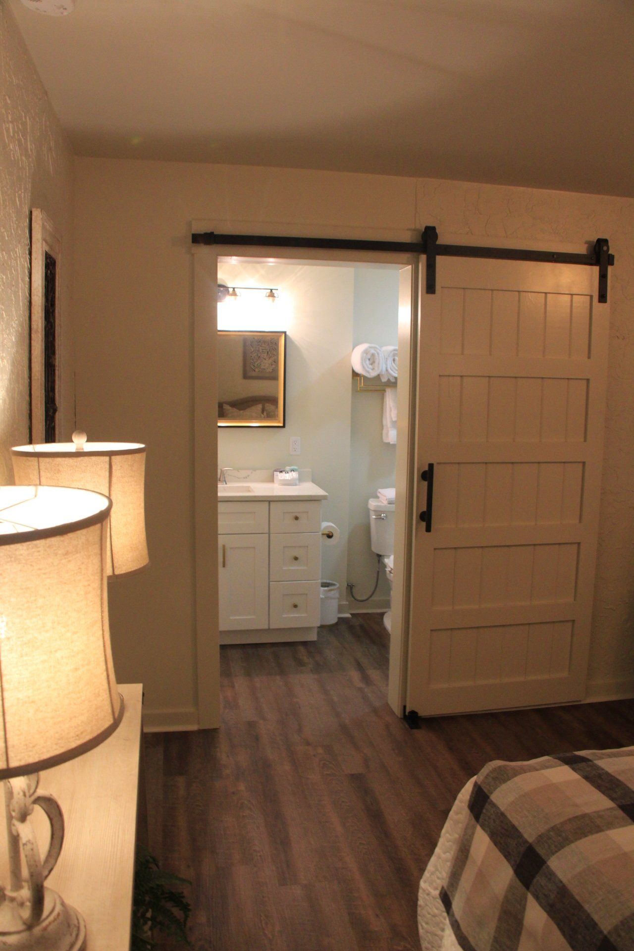 Bedroom with a sliding barn door leading to a bathroom. White door and vanity, dark wood floors, two lamps, and a bed.