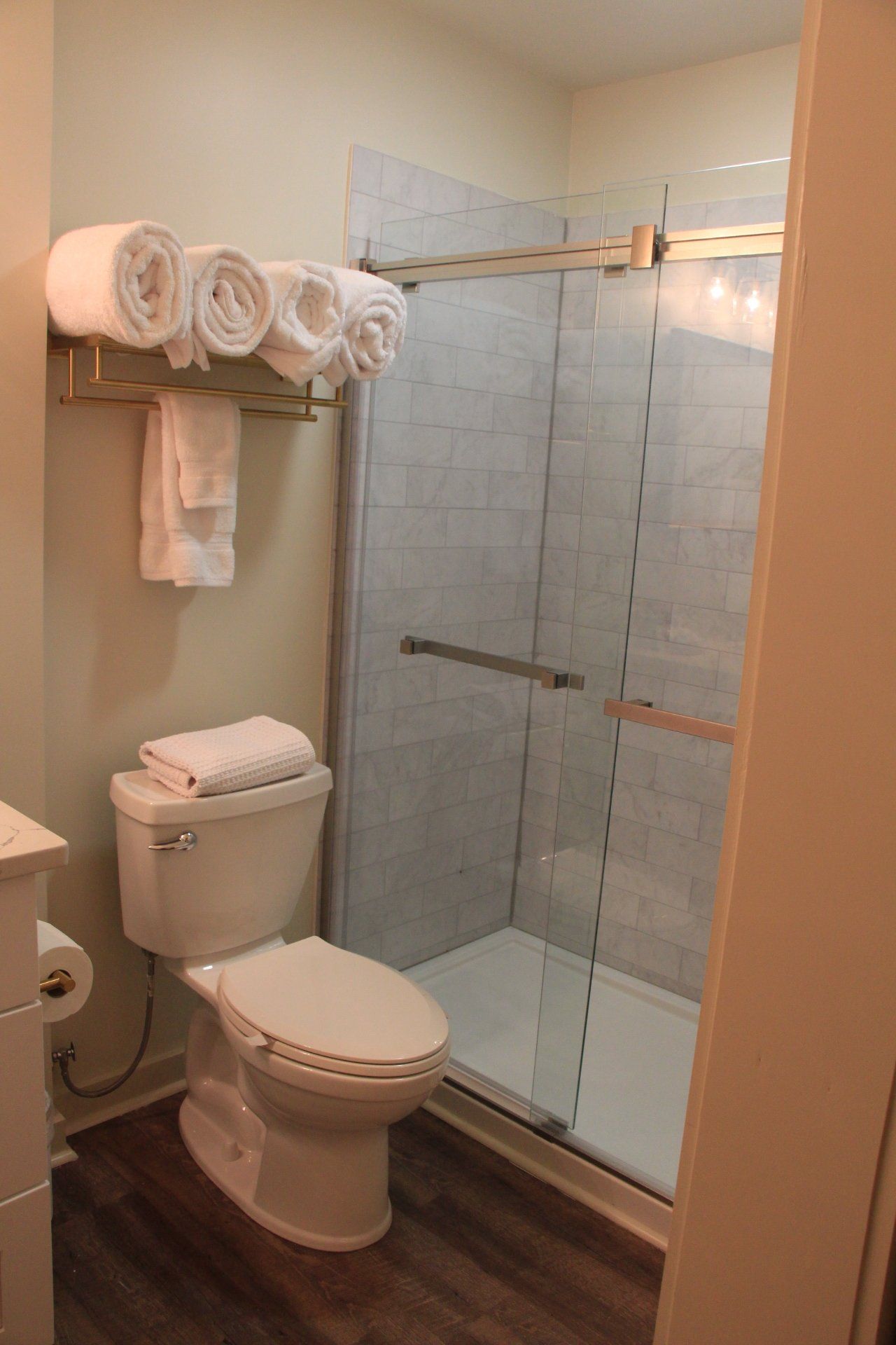 Bathroom with a toilet, shower with glass doors, and towels on a rack. The floor is wood-like.
