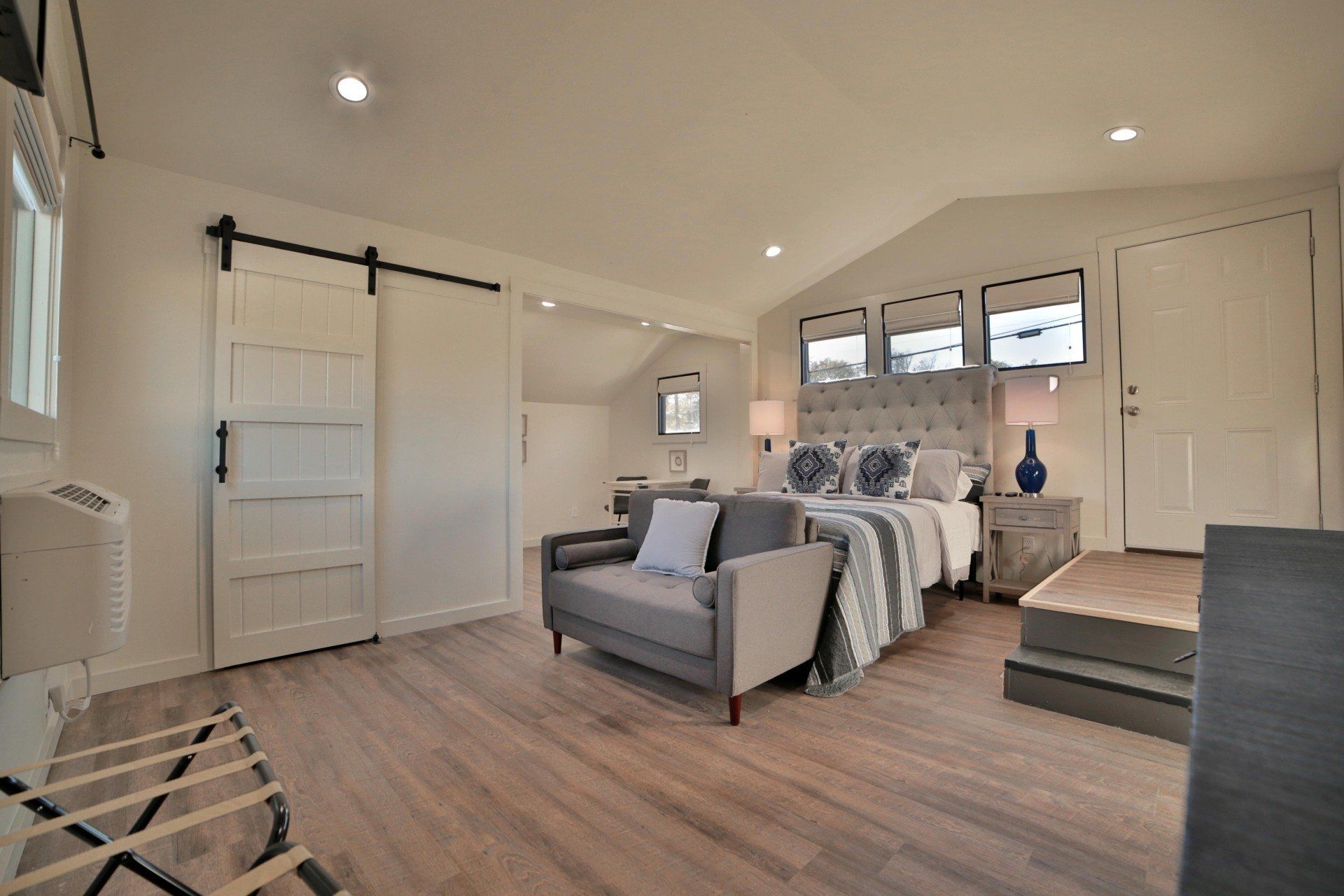Bedroom with a king-sized bed, gray sofa, white barn door, and wood floors.