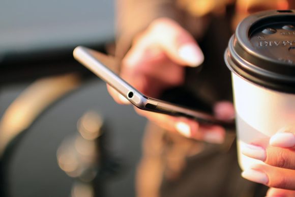 Person holding a smartphone and a coffee cup. The phone is black, and the cup is white with a black lid.