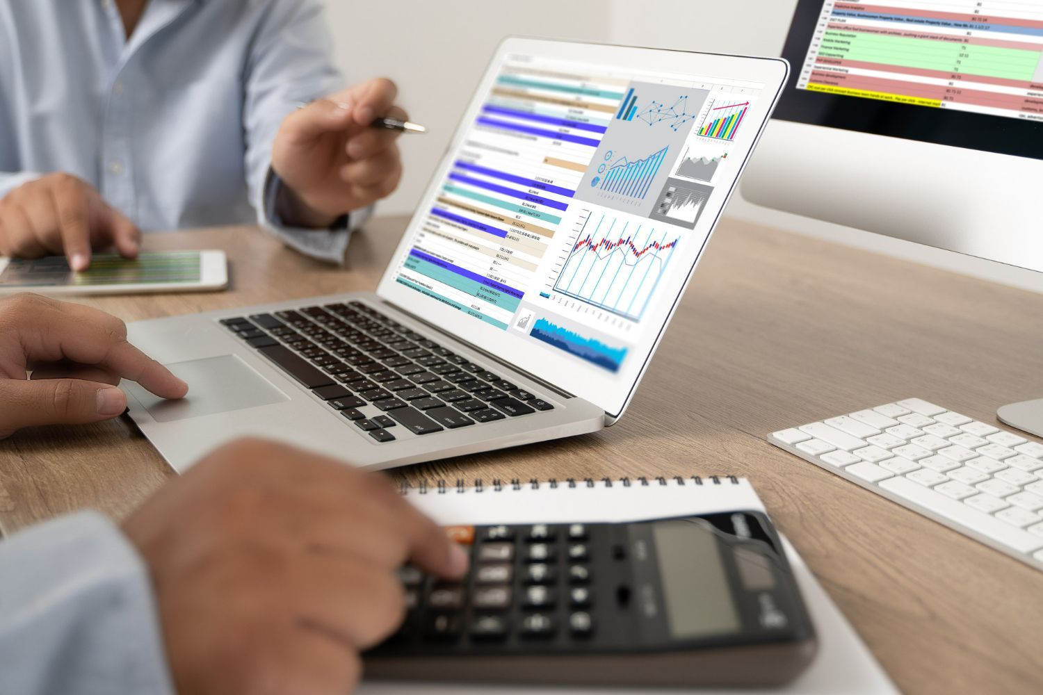 People reviewing financial data on a laptop and monitor, using a calculator and notepad.