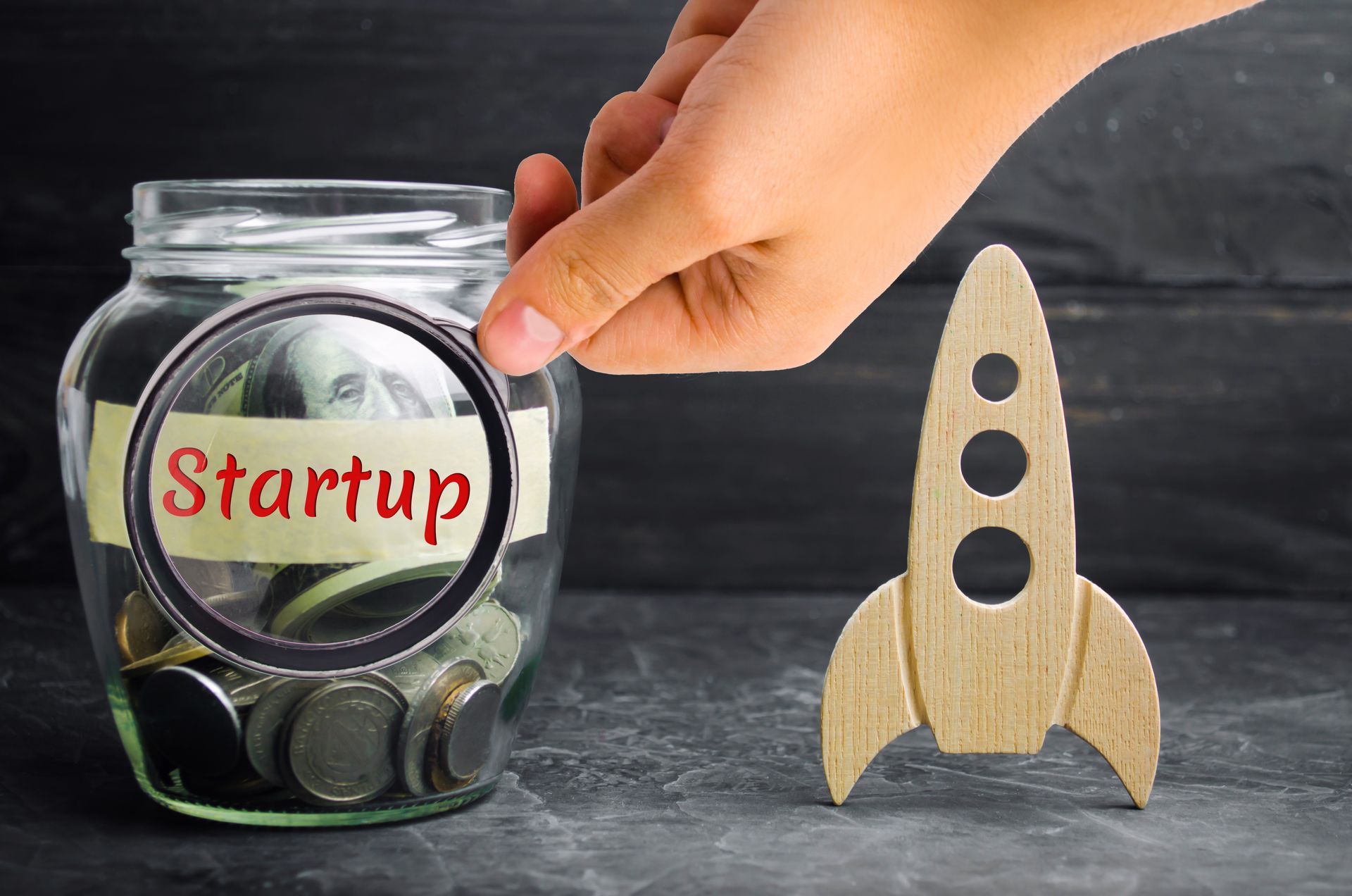 Hand placing a coin in a jar labeled “Startup” beside a small wooden rocket