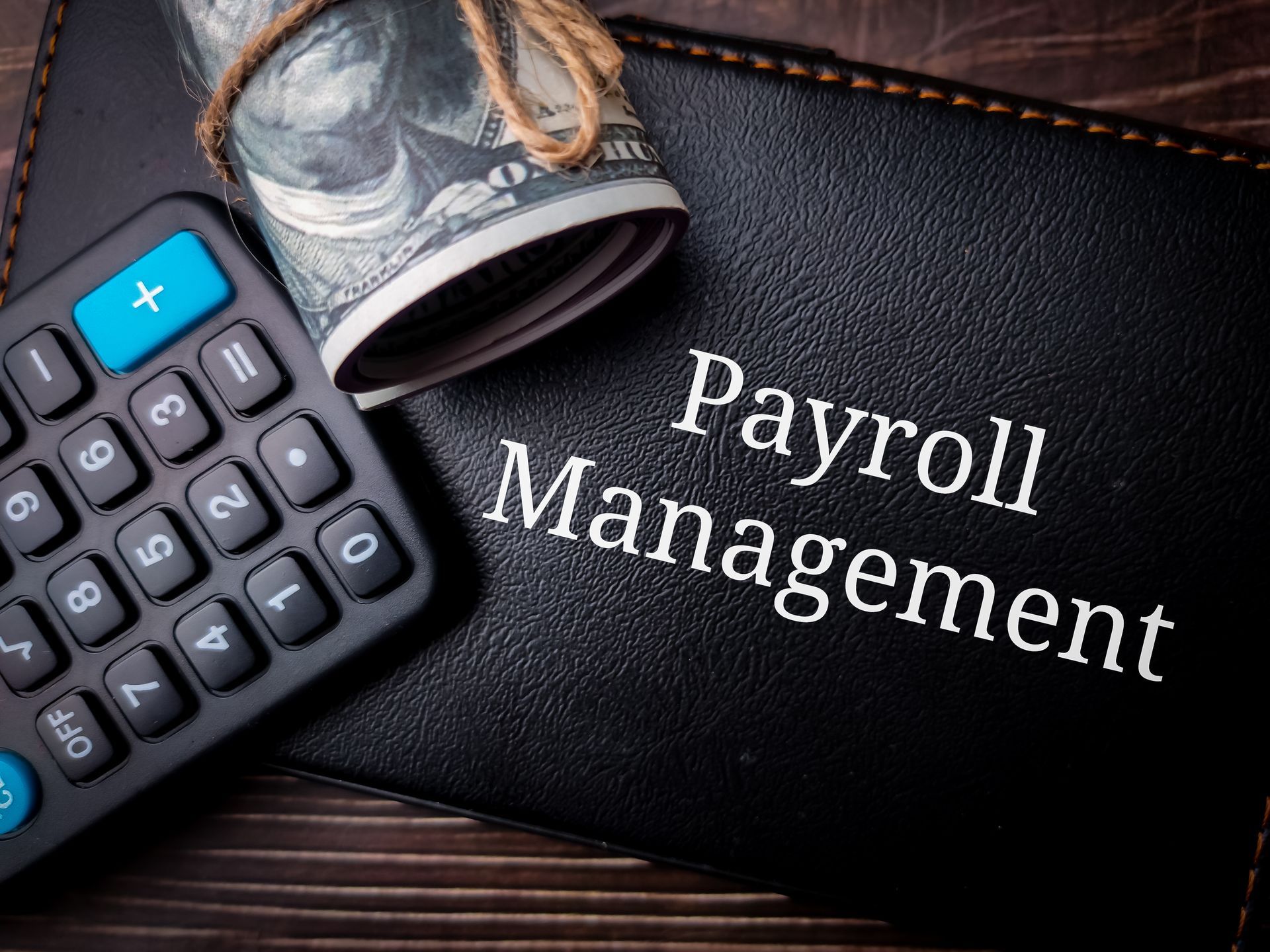 Calculator beside rolled cash on a black notebook labeled “Payroll Management”