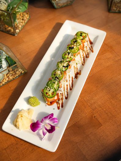Sushi roll topped with sliced jalapeños and sauce on a white plate, garnished with orchid and wasabi.