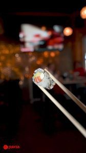 Two chopsticks holding a bite of sushi over a blurred restaurant table and lights