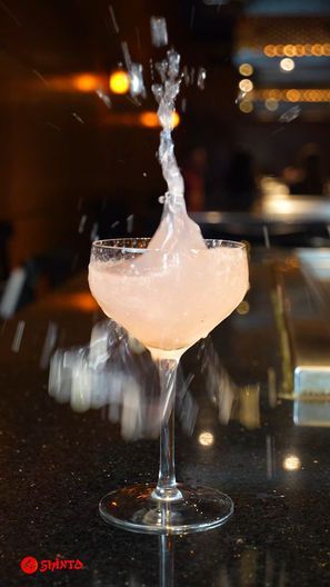 Cocktail glass with a drink splashing upward on a bar counter, warm lights blurred in the background