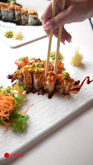 Chopsticks picking up a glazed sushi roll on a white plate with greens and shredded carrot