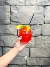 Hand holding a red cocktail with lemon and lime garnish, black straw, against a stone wall.